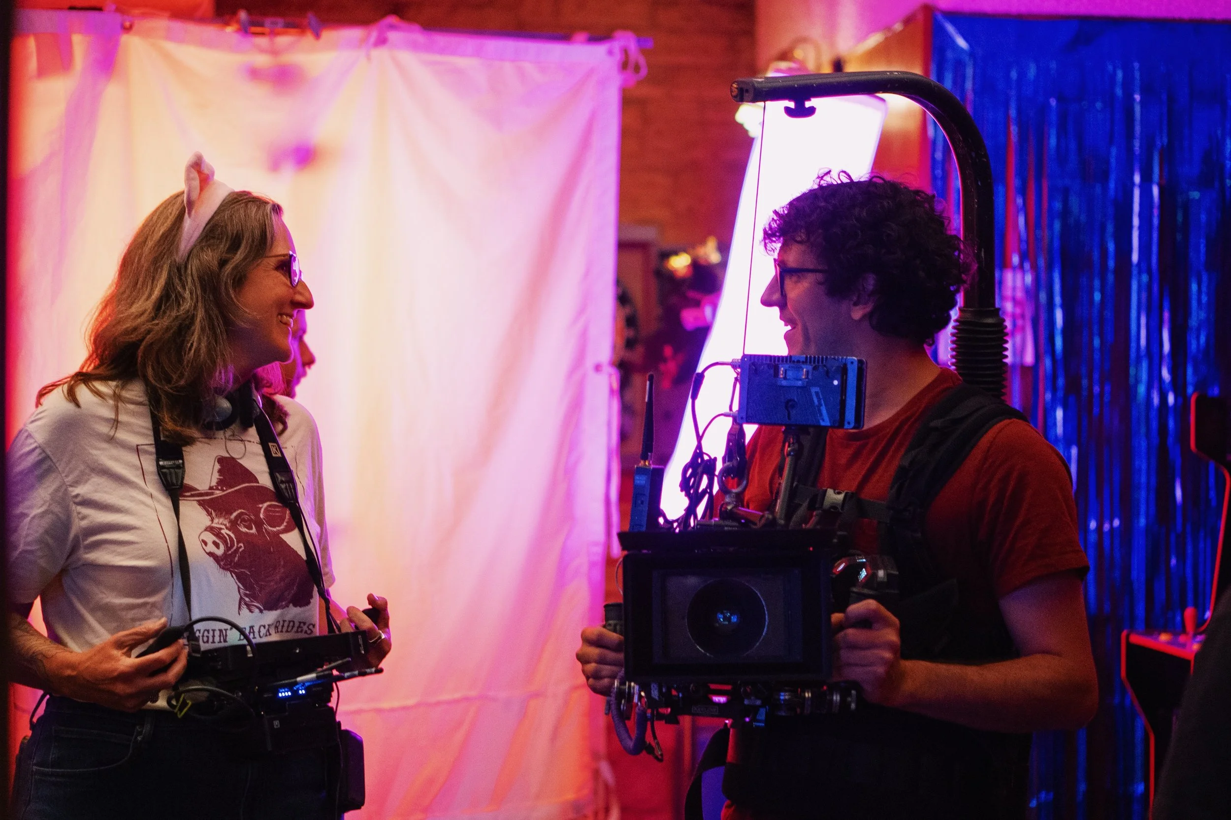 A filmmaker wearing a short and a headband, smiling and looking right towards a camera operator holding a camera. They are in front of a white sheet/background