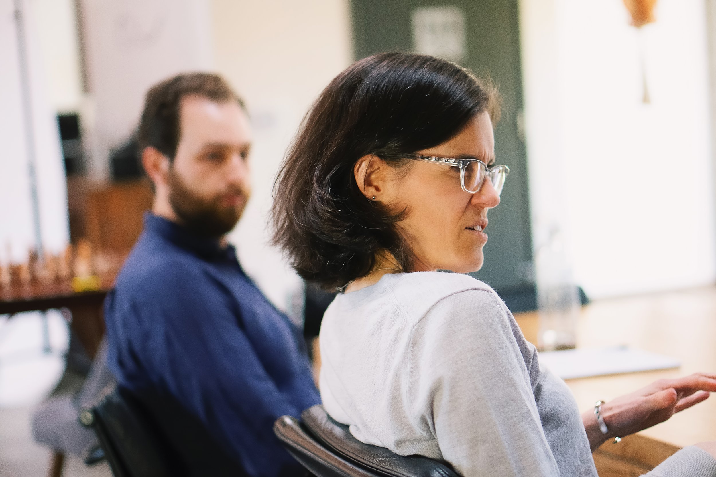 A white woman with glasses and a great long-sleeved shirt sits at a table, engaged in conversation. A white man with a beard in a blue shirt looks on in the background.