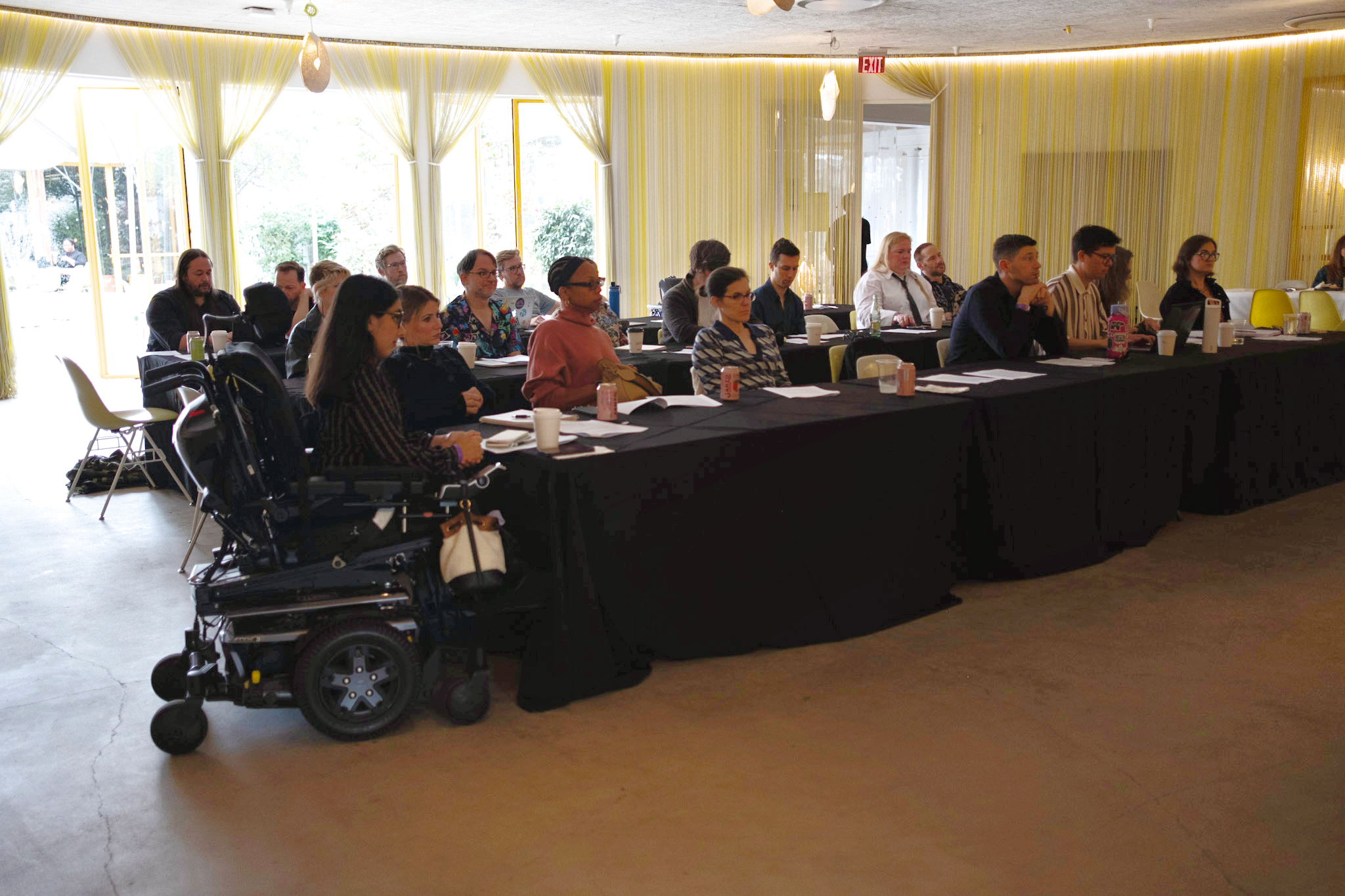  People sit attentively at a long, black-covered table in a well-lit room with sheer curtains. A person in a wheelchair is visible in the foreground. 