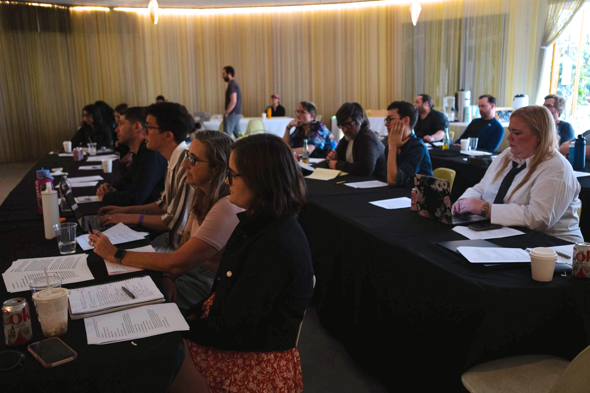  A group of people sit in a dimly lit conference room, intently focused on a presentation. They have laptops, papers, and drinks on black tables. 