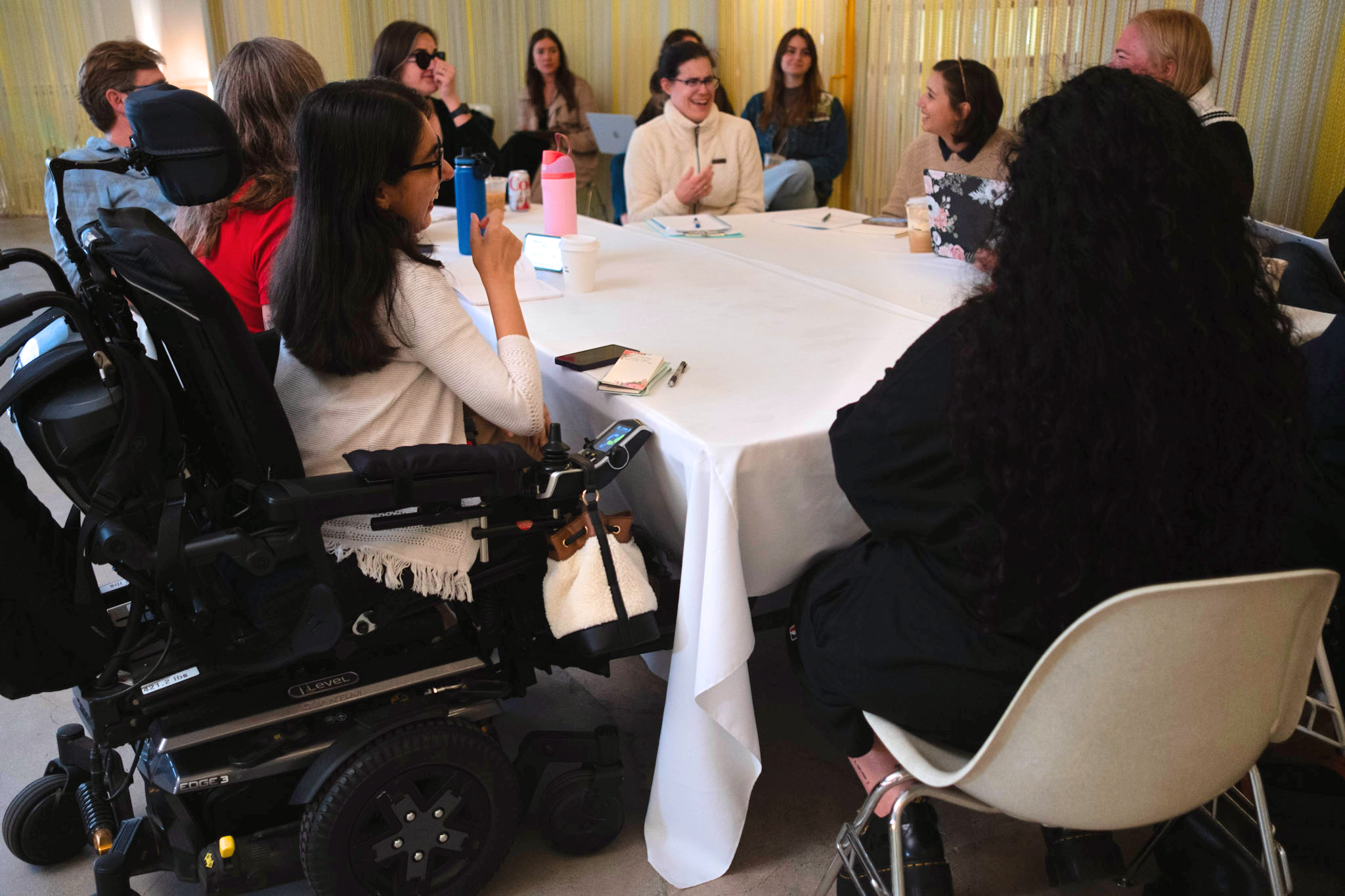  A group of people sit around a large, white, round table in a casual meeting. One person is in a wheelchair, visible on the left. The atmosphere is engaged and collaborative. 