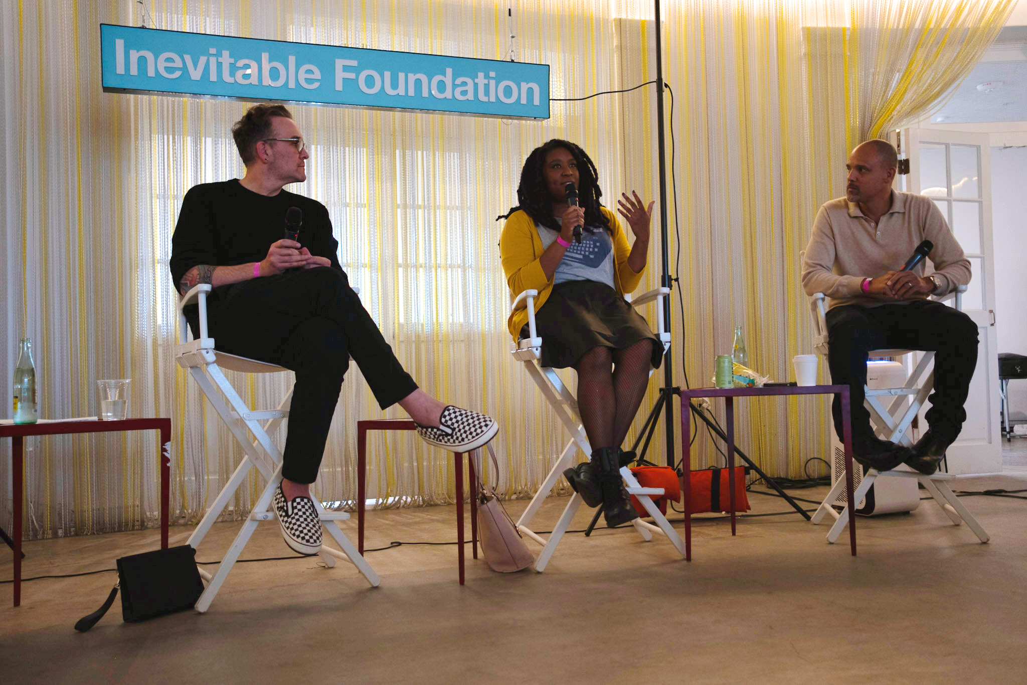 Three people sit in director’s chairs in front of an "Inevitable Foundation" sign as part of a panel discussion. The middle person, who is a Black woman with braids, speaks into a microphone.
