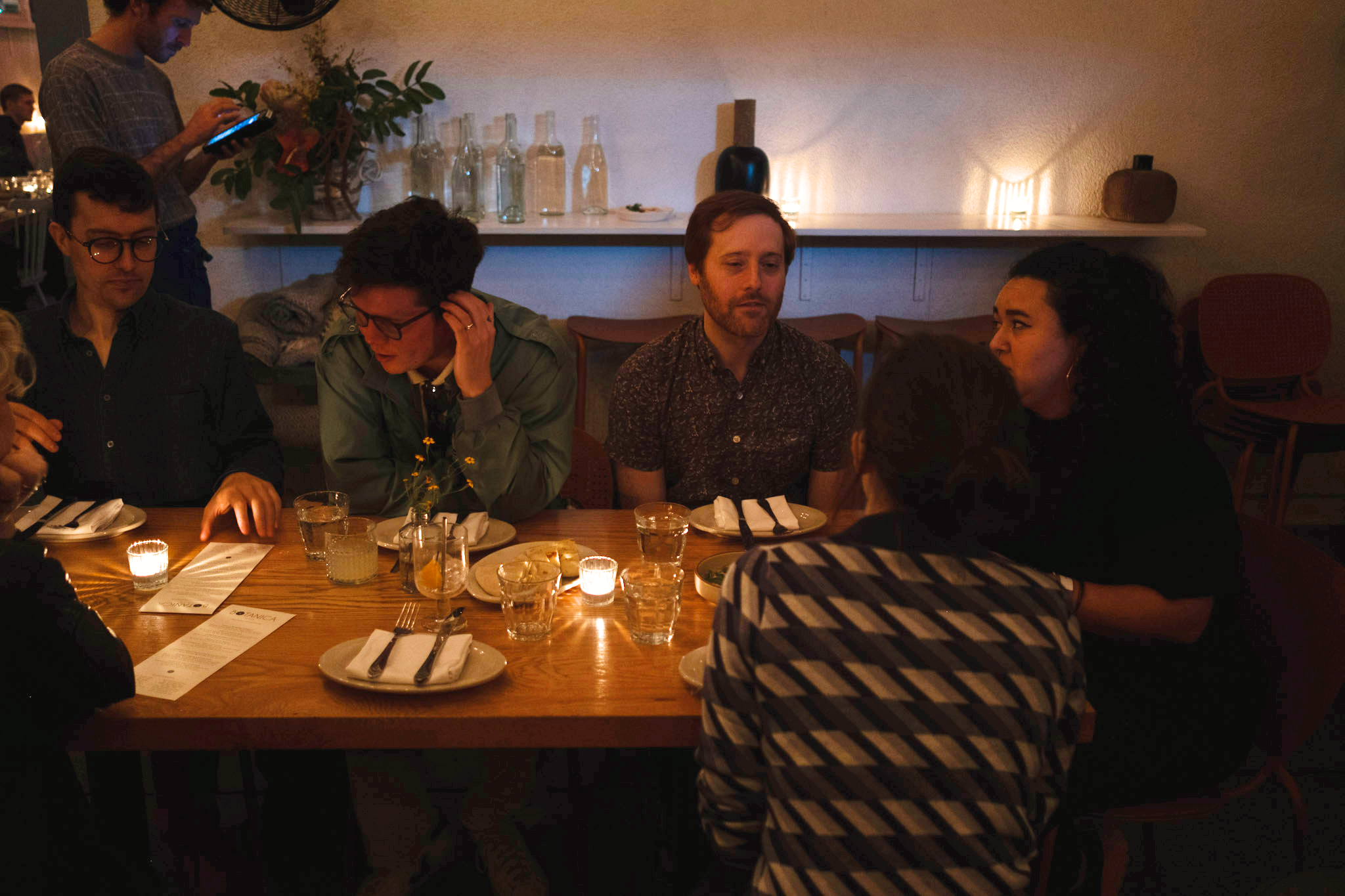  A cozy dinner setting shows six people seated at a wooden table with glasses, plates, and menus. Warm candlelight creates an intimate atmosphere. 