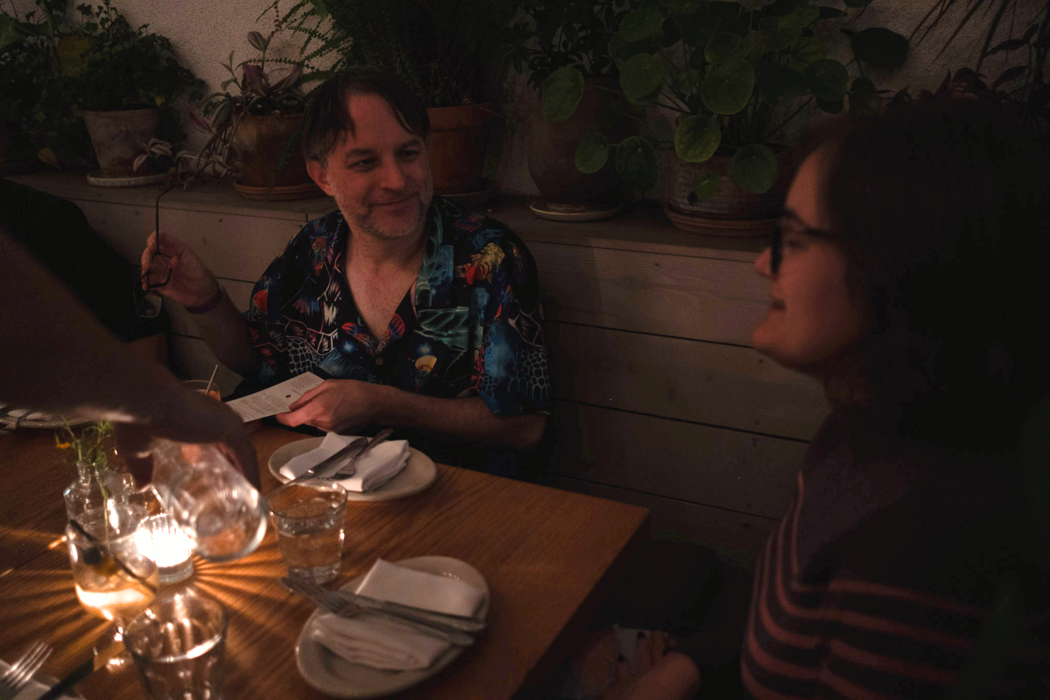  A warmly lit restaurant scene with two people at a wooden table. The man, smiling, holds a menu near potted plants. Glasses and plates are neatly arranged, creating a cozy ambiance. 