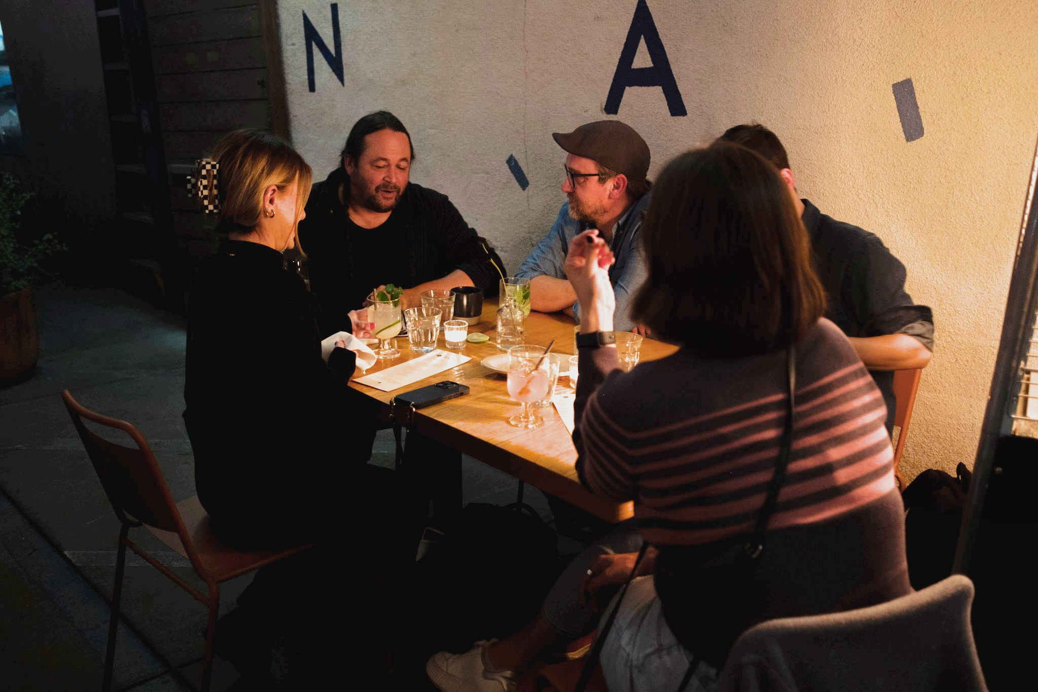  A group of five people is seated at a dimly lit table, engaged in conversation. Glasses and a menu are on the table, creating a relaxed atmosphere. 