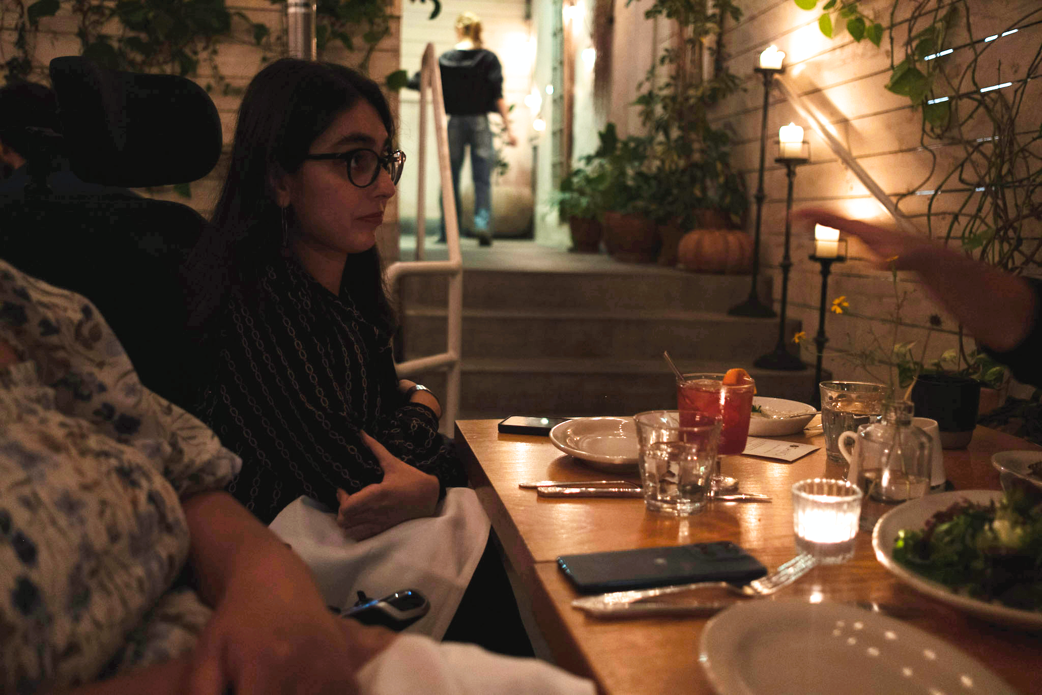  A woman with glasses sits in her wheelchair at a dimly-lit, candlelit table in a cozy restaurant. She is listening intently. A person walks up steps in the background. 