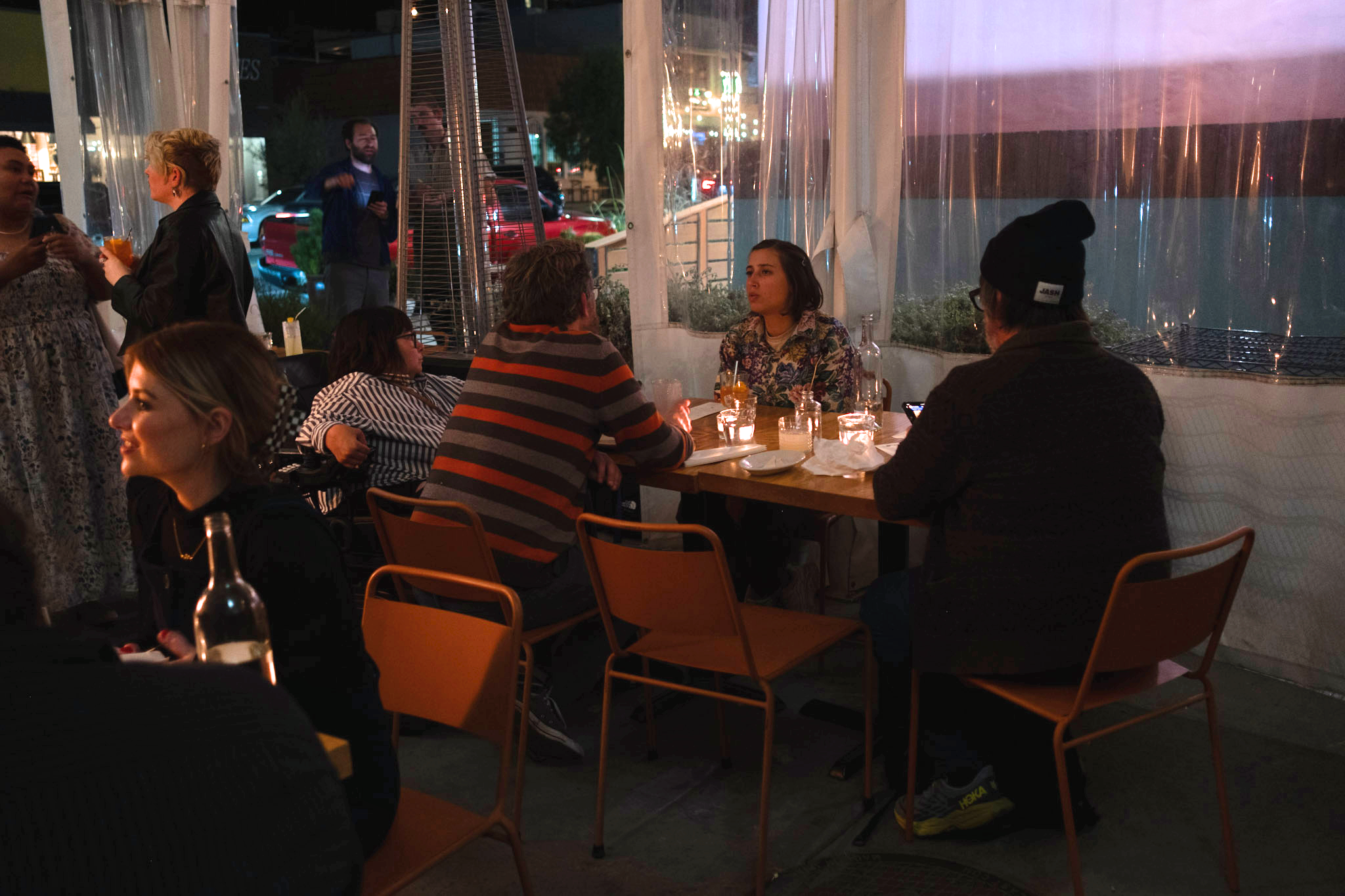  A cozy outdoor restaurant scene at night. Five people sit at tables under warm candlelight, while others chat nearby. The atmosphere is relaxed and intimate. 