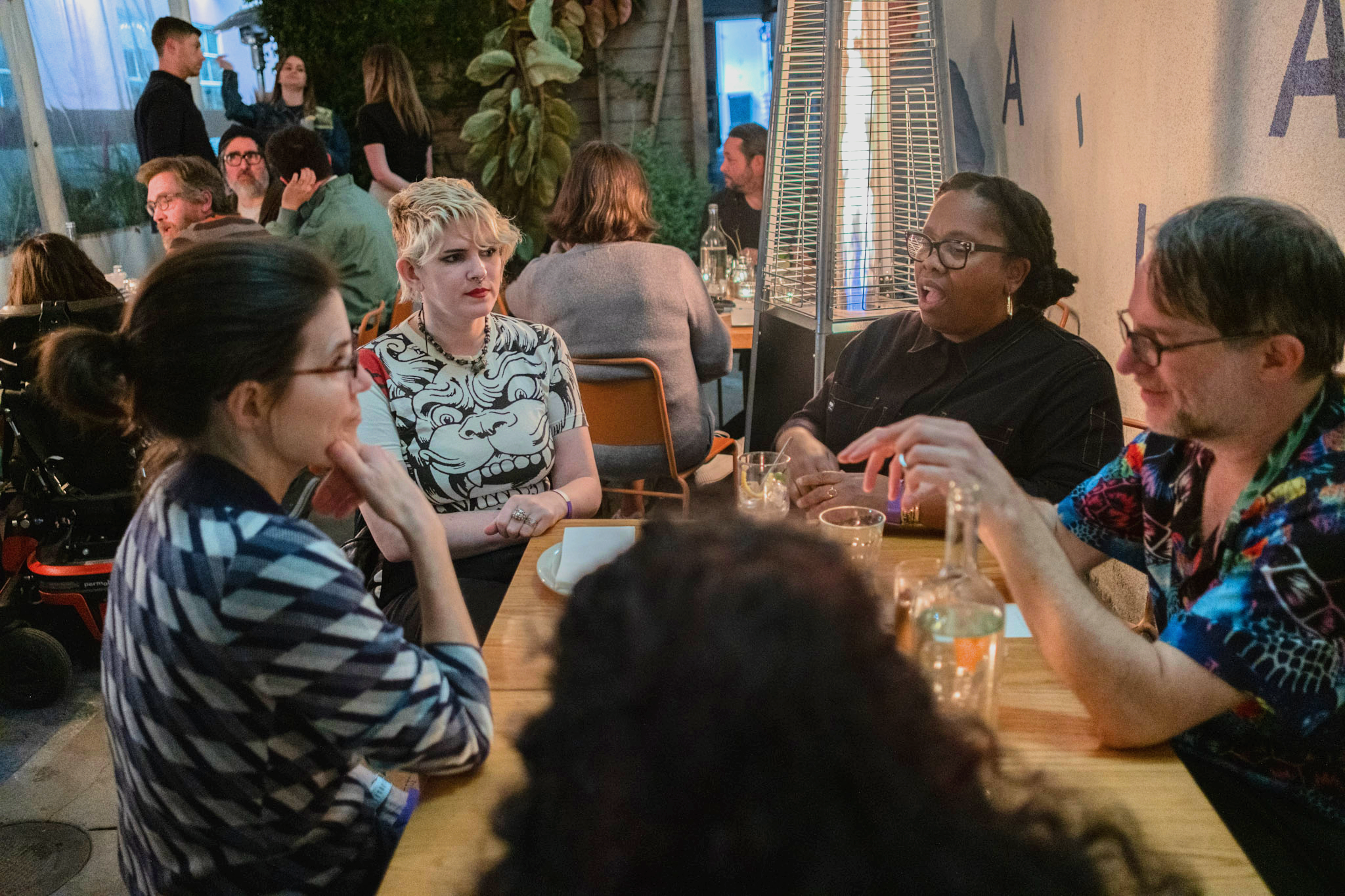  A group of people sit around a wooden table in a lively restaurant listening to each other intently. There is an ASL interpreter at one end of the table. A heater glows nearby. 