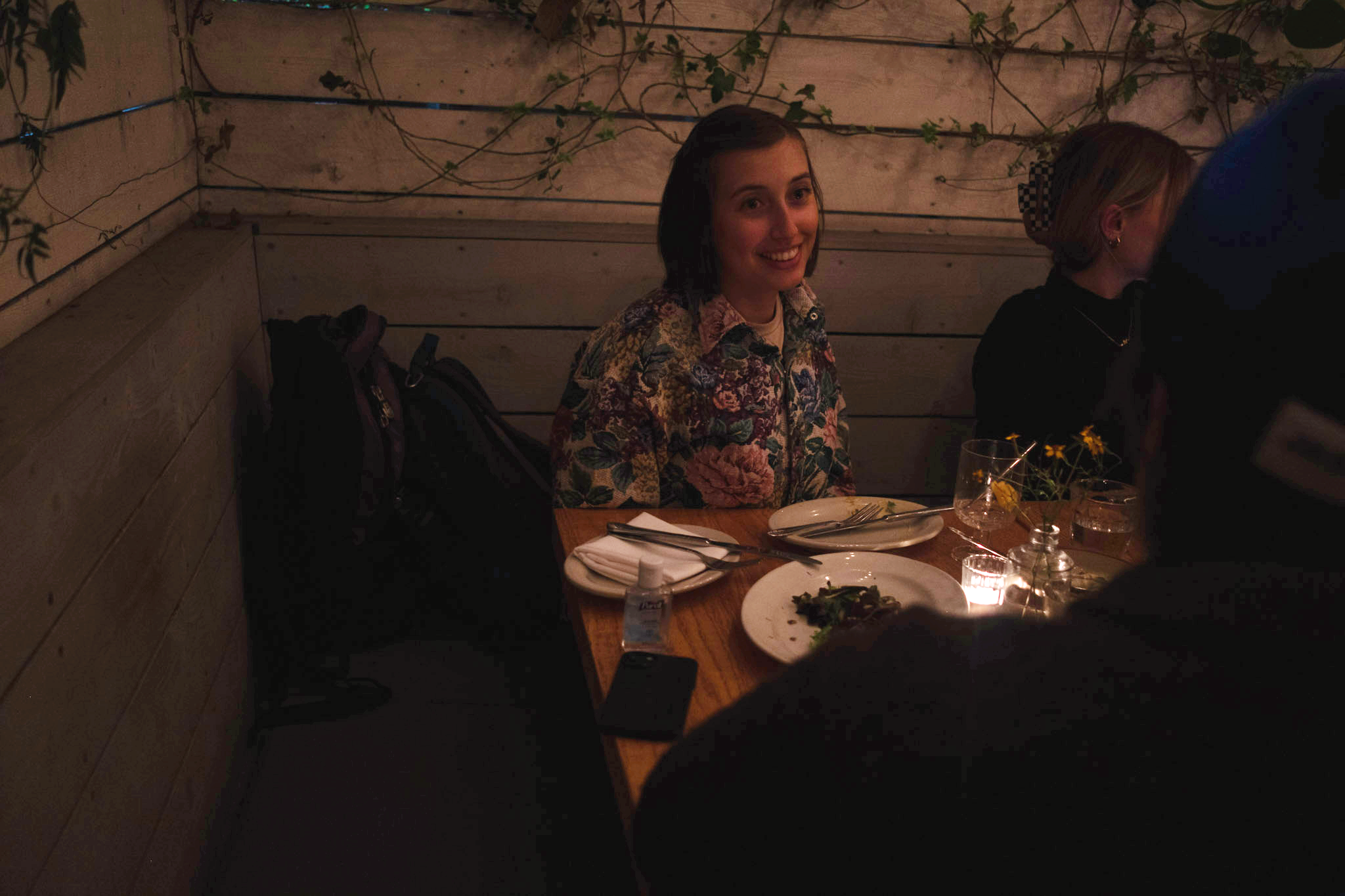 Smiling woman in a floral shirt sits at a candle-lit table with friends. Behind her hang plants on a wooden wall. 