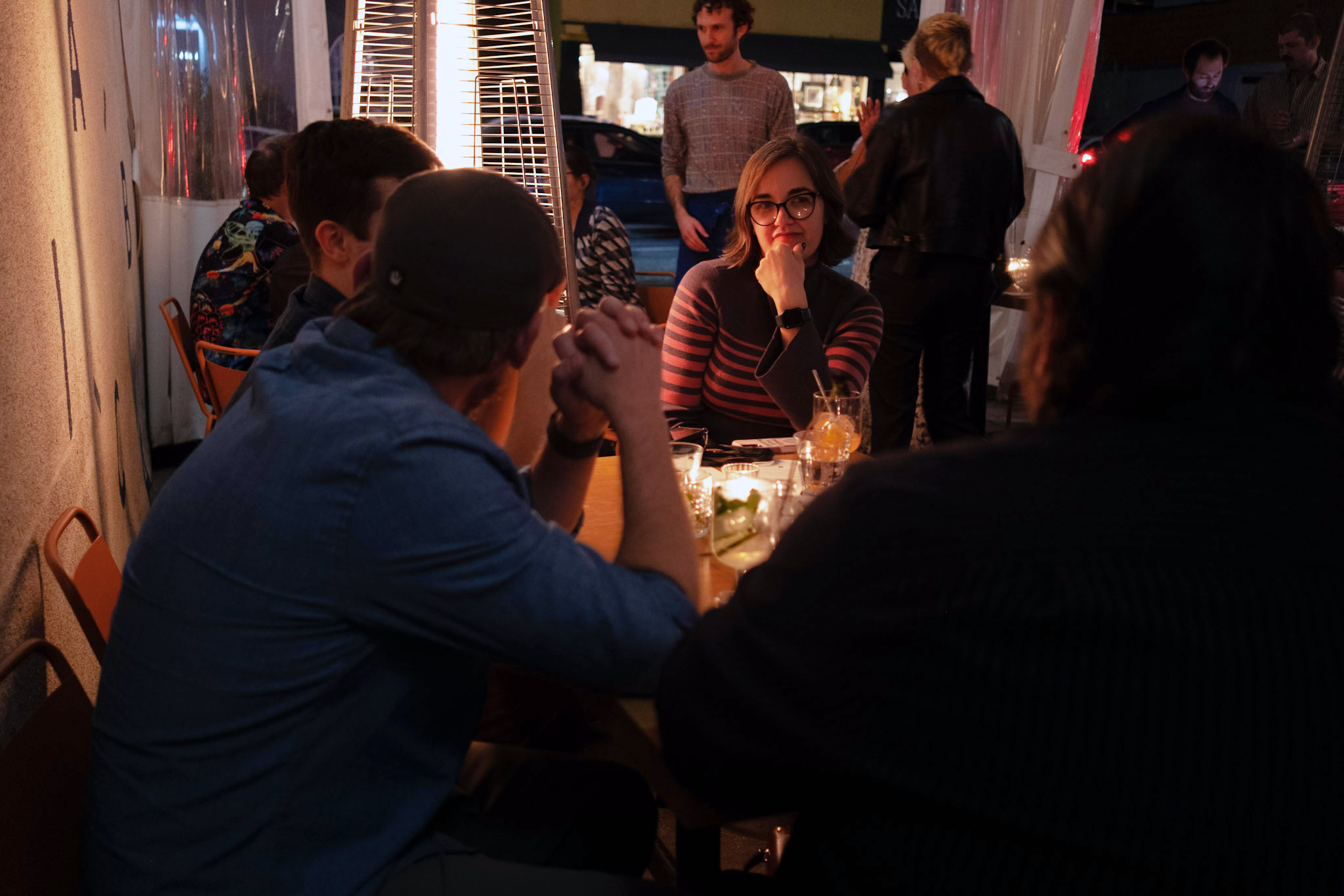  Outdoor evening gathering with screenwriters around a table, which is lit by a tall heater. A person in a striped sweater listens intently, setting a warm, intimate tone. 