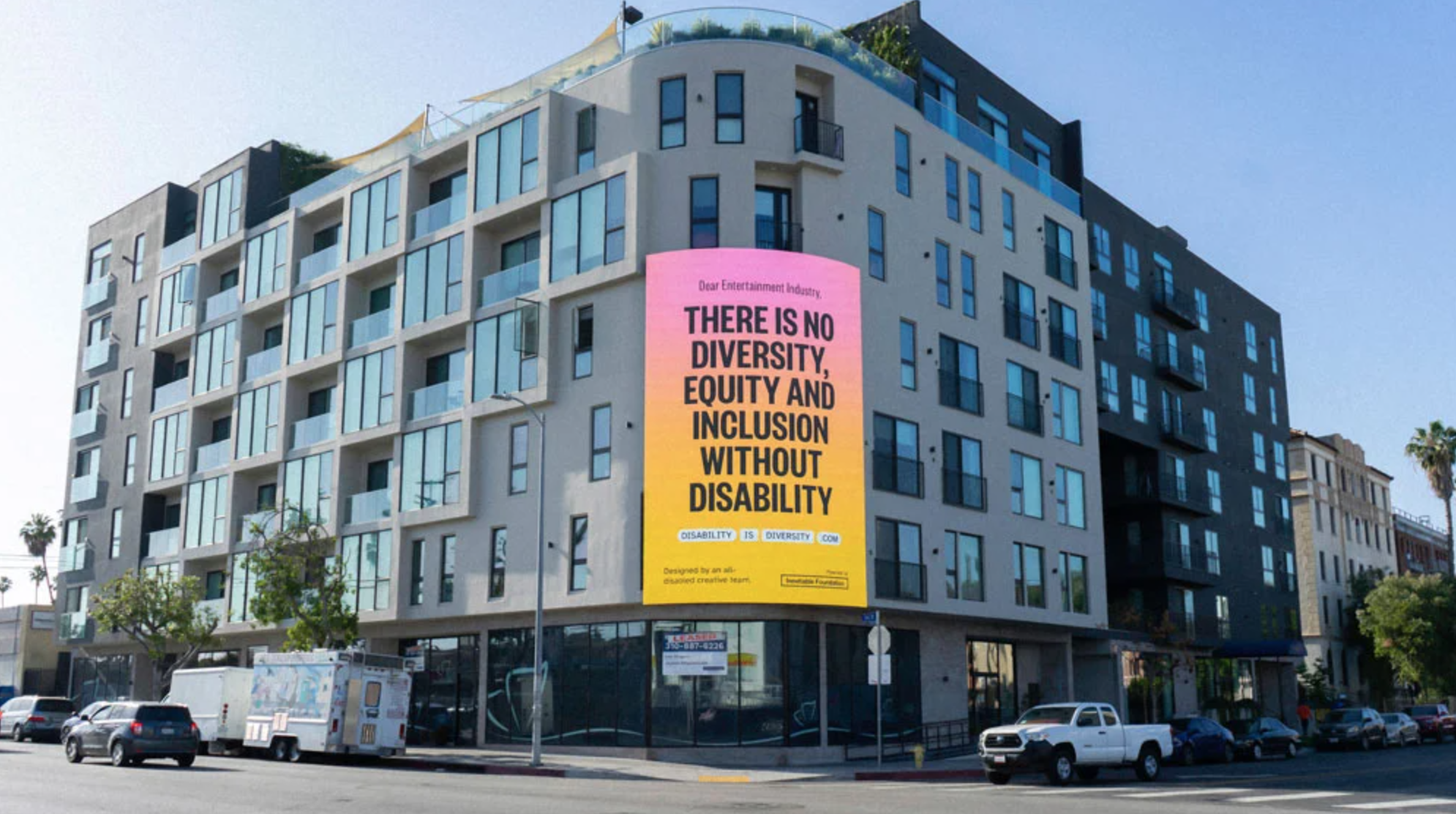 Modern apartment building on a street corner with a large pink and yellow banner that  reads there is no diversity, equity and inclusion without disability in bold letters. Several cars are parked on the street nearby.