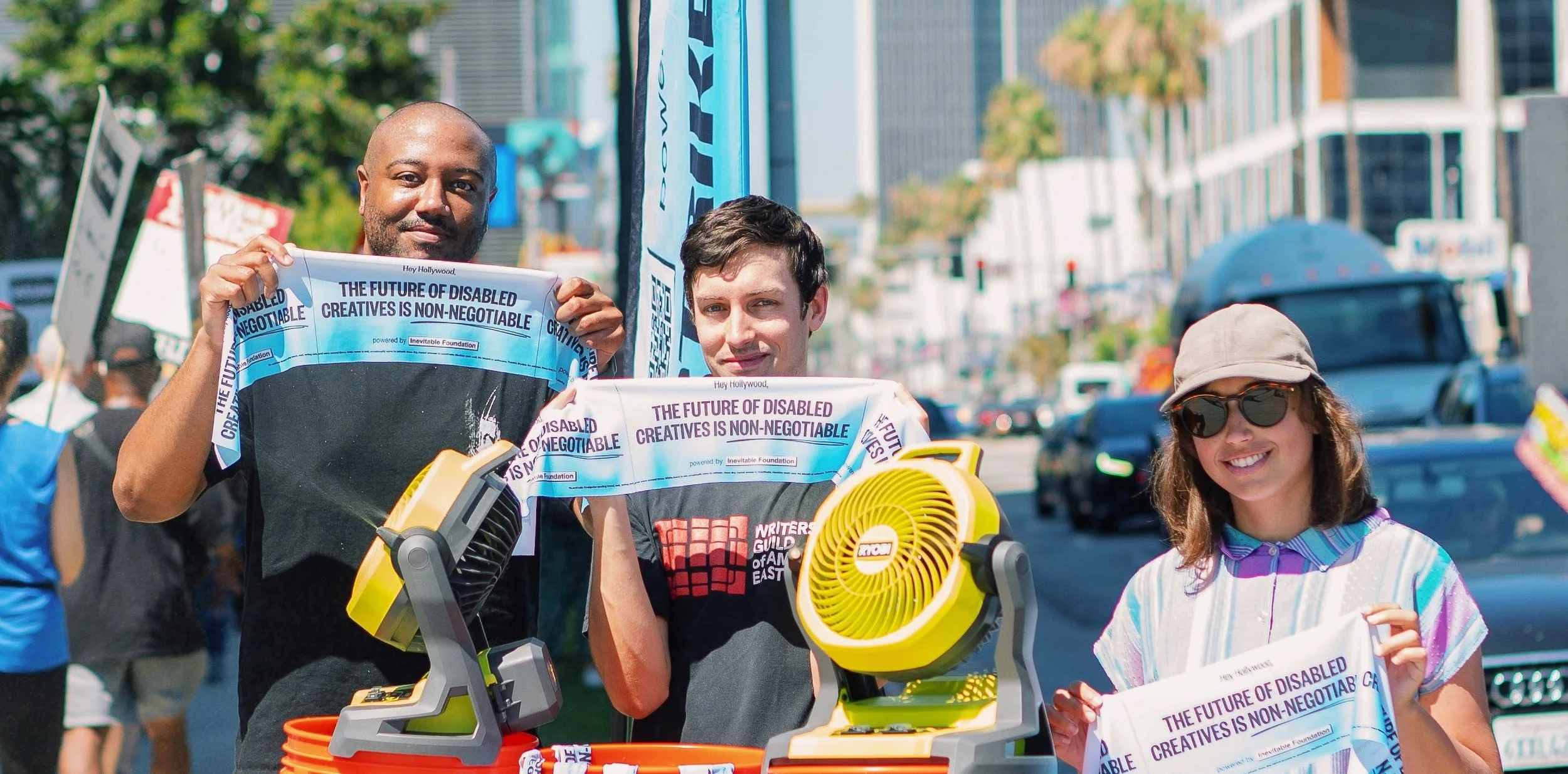 One Black man, one white man and one Latina  woman stands outdoors holding banners that read, The Future of Disabled Creatives is Non-Negotiable. They smile at the camera, with street lined with palm trees and buildings.