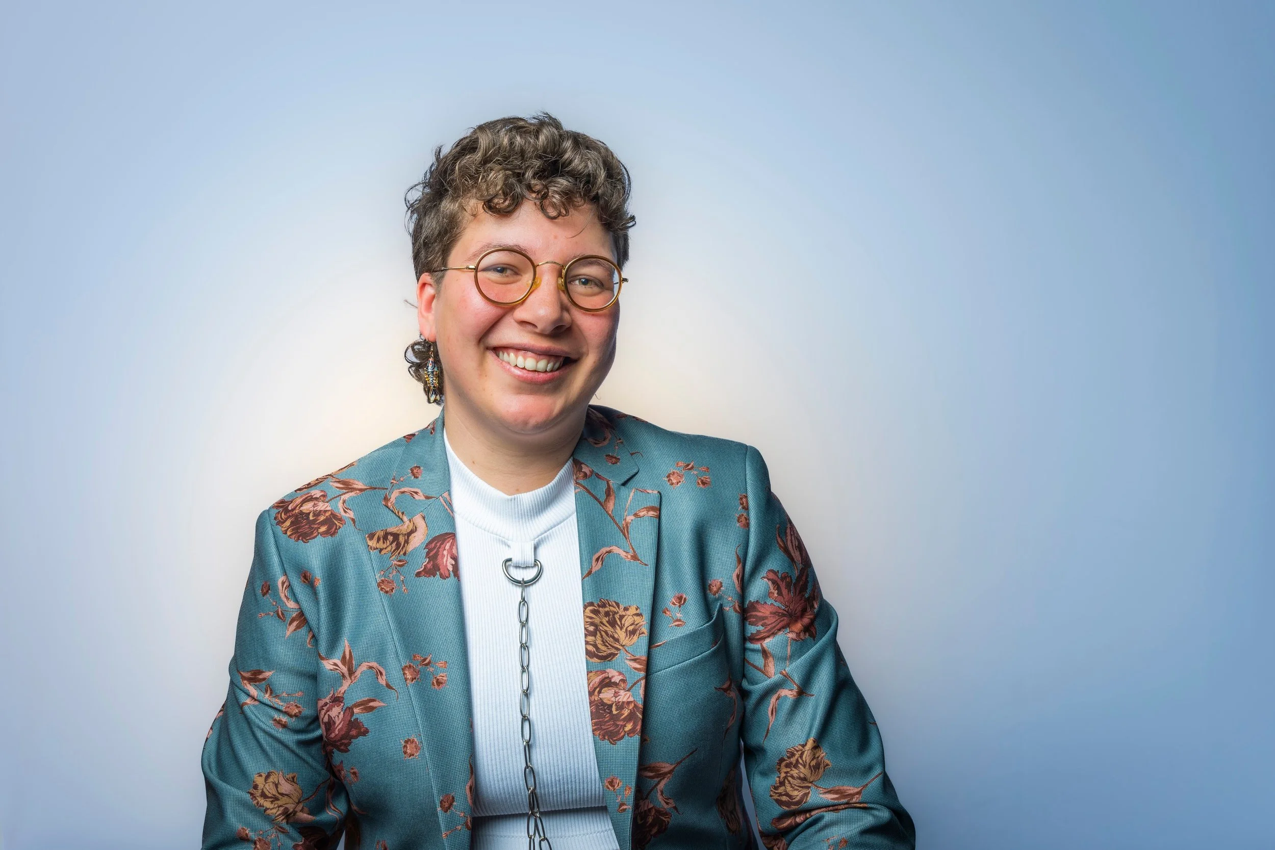 A 30-year old Dutch-Indonesian nonbinary person with a short brown curly mullet and a round face, smiling widely.
