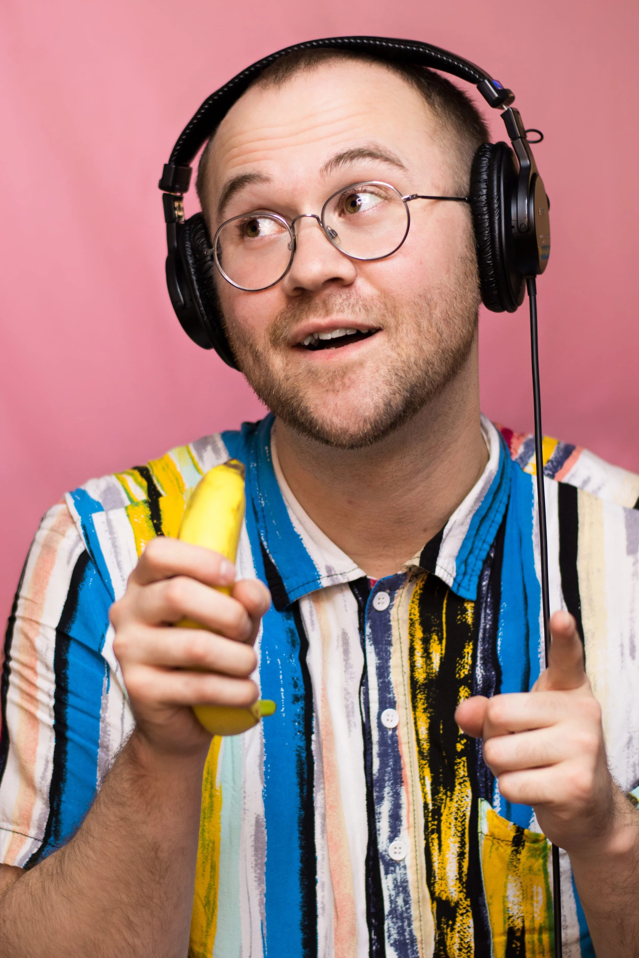A 30 year old white man with buzzed brown hair and brown eyes, smiling and looking off to the left while pretending to speak into the banana in his hand like it's a microphone. He is wearing wire frame glasses.