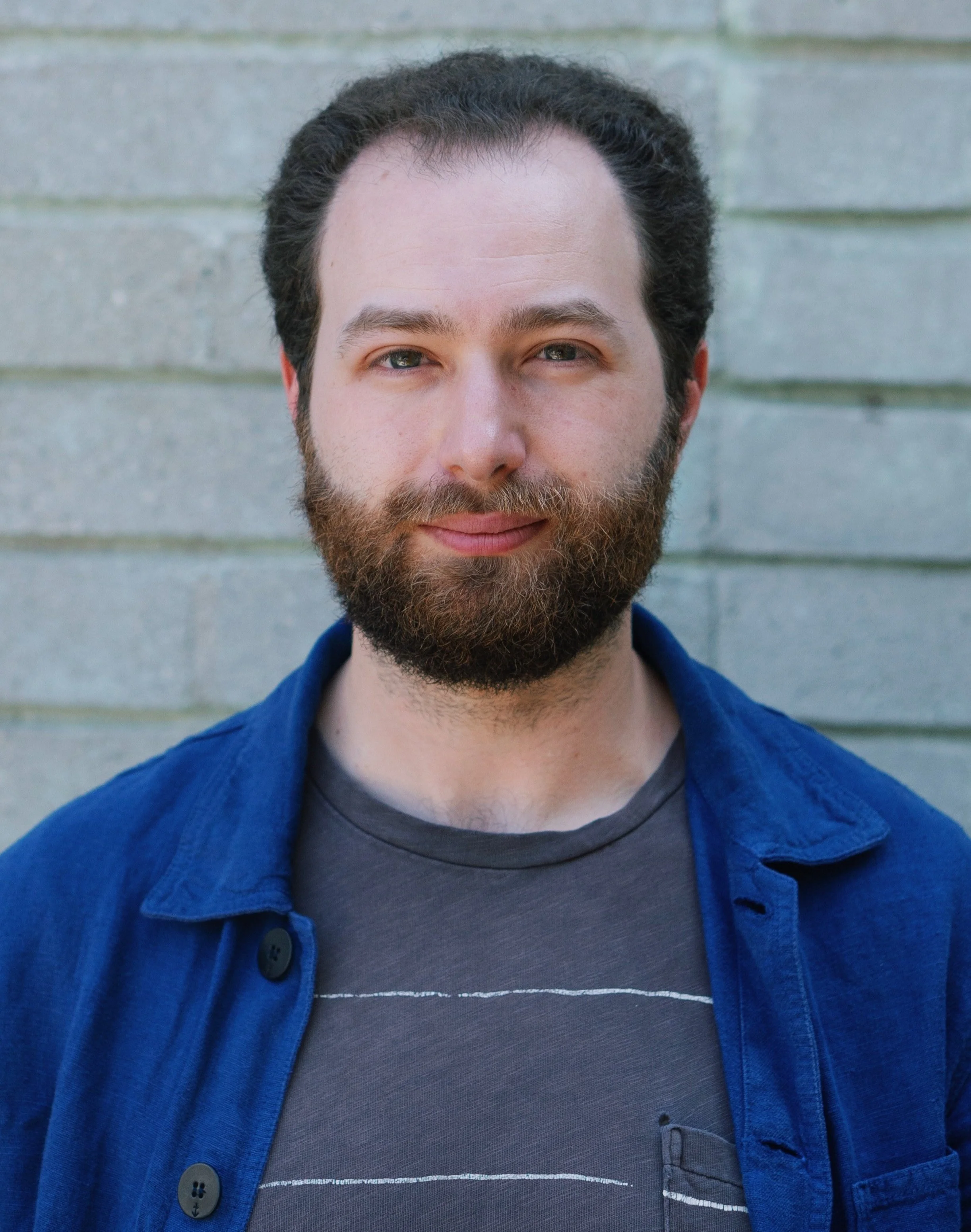 Headshot of a white man with dark brown hair and a bushy brown beard. He has hazel eyes and is smiling slightly at the camera. He's wearing a dark grey t-shirt and a blue jacket.