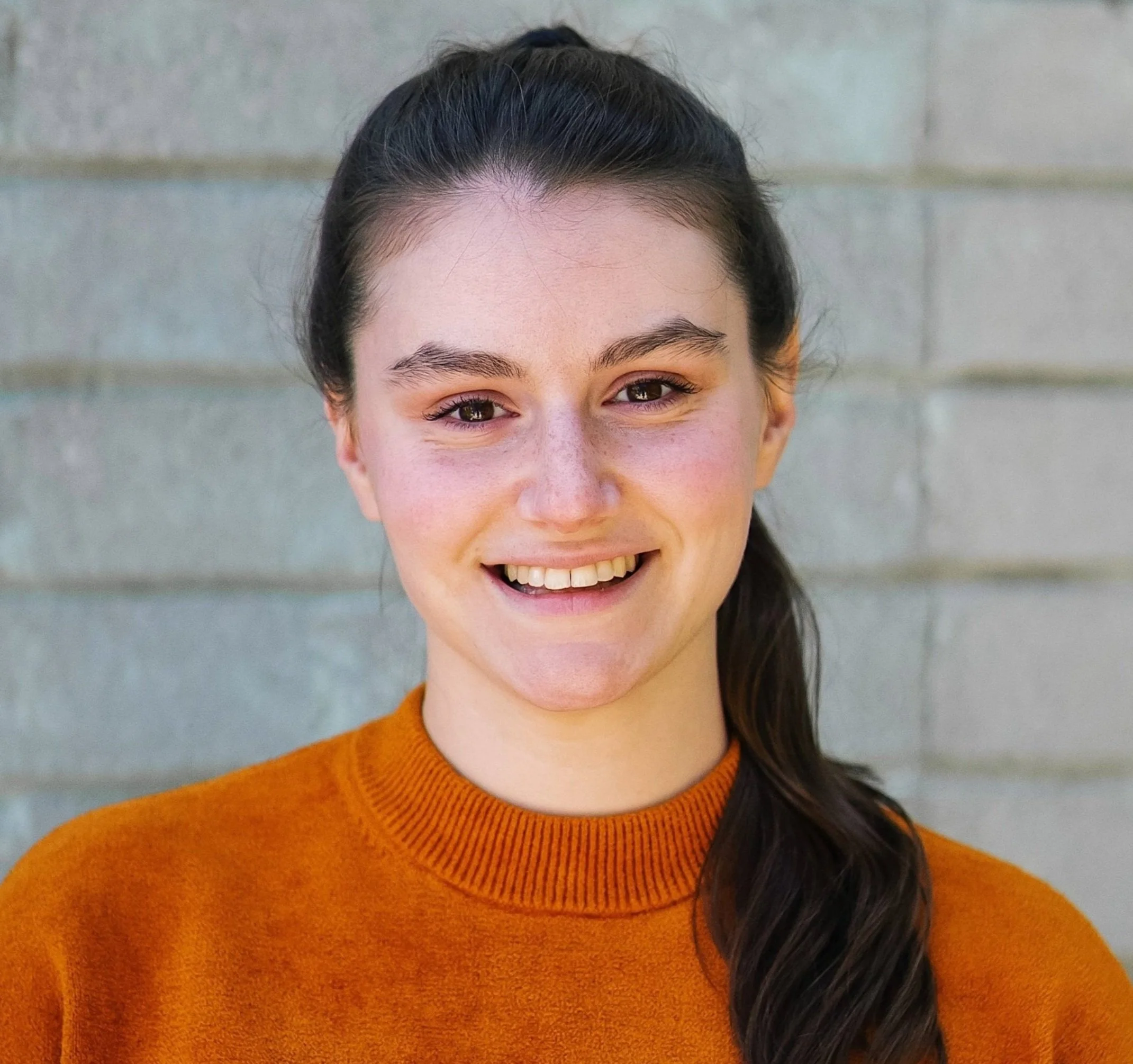 Headshot of a white woman with dark brown hair pulled back in a long ponytail. She has brown eyes and is smiling at the camera. She's wearing a velvety orange sweatshirt.