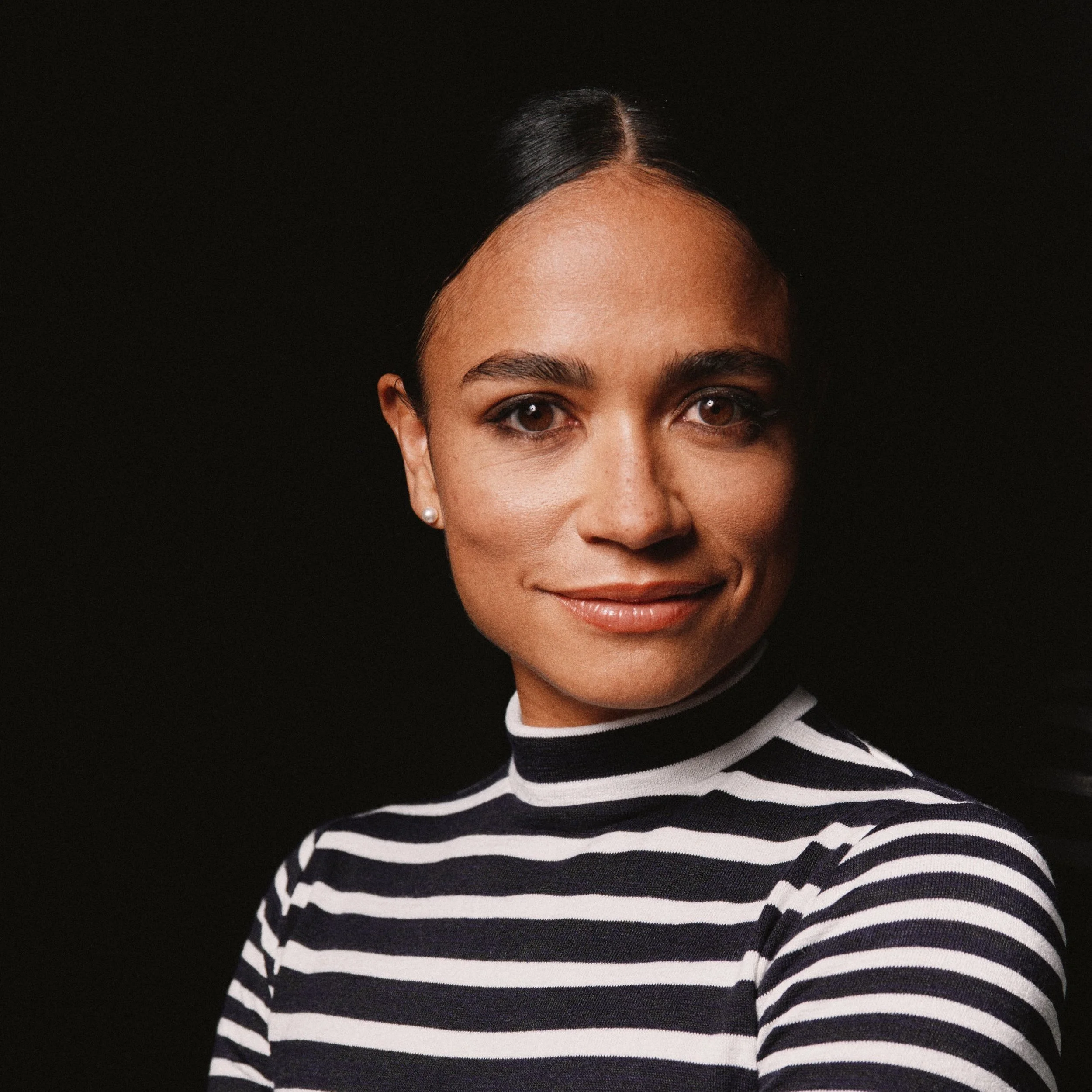 A Black woman in a black and white striped mock neck shirt. She looks into the camera, smiling.
