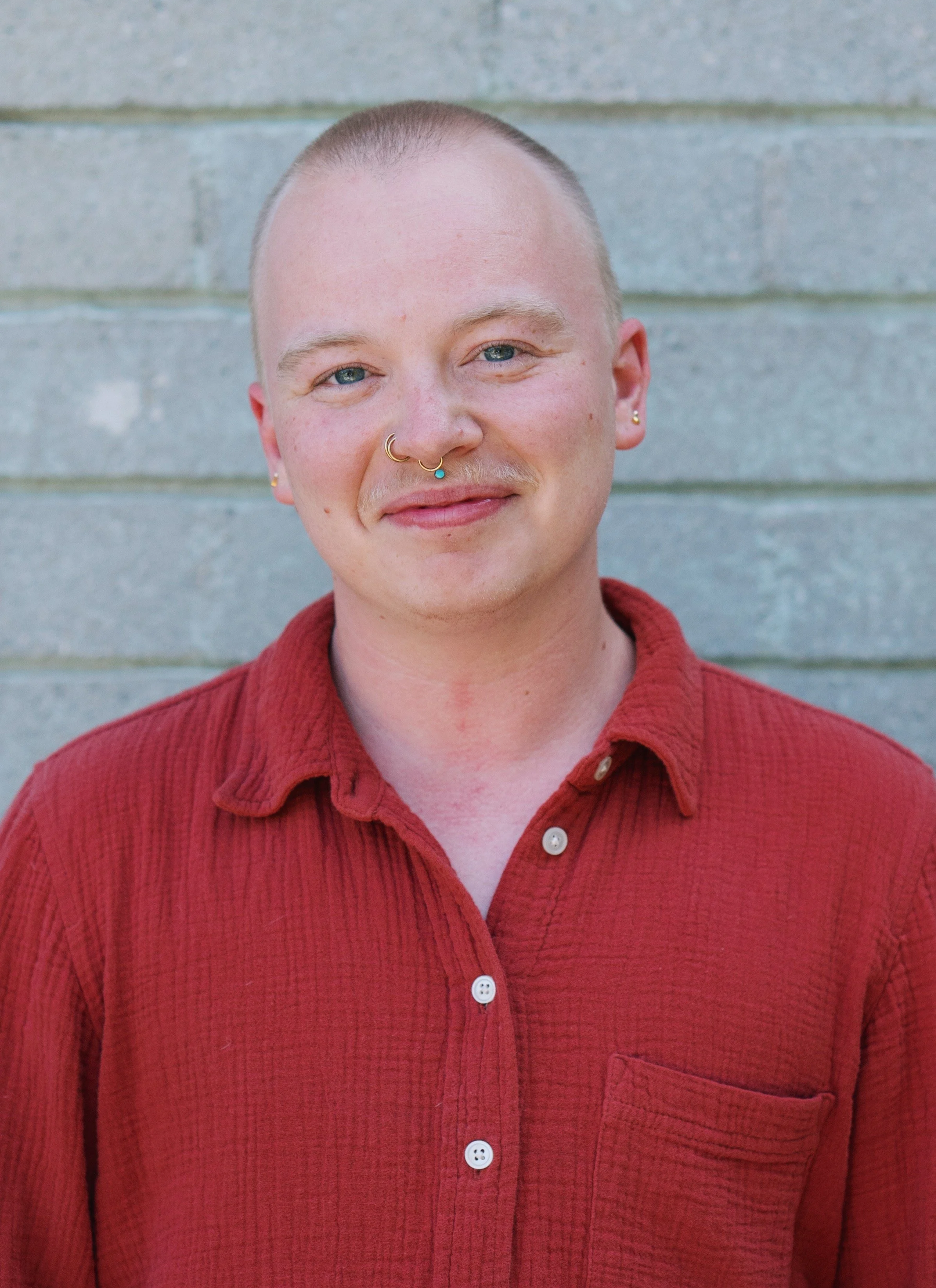A white person with a shaved head, wearing a red linen collared shirt. Their blue eyes match their turquoise facial jewlery.