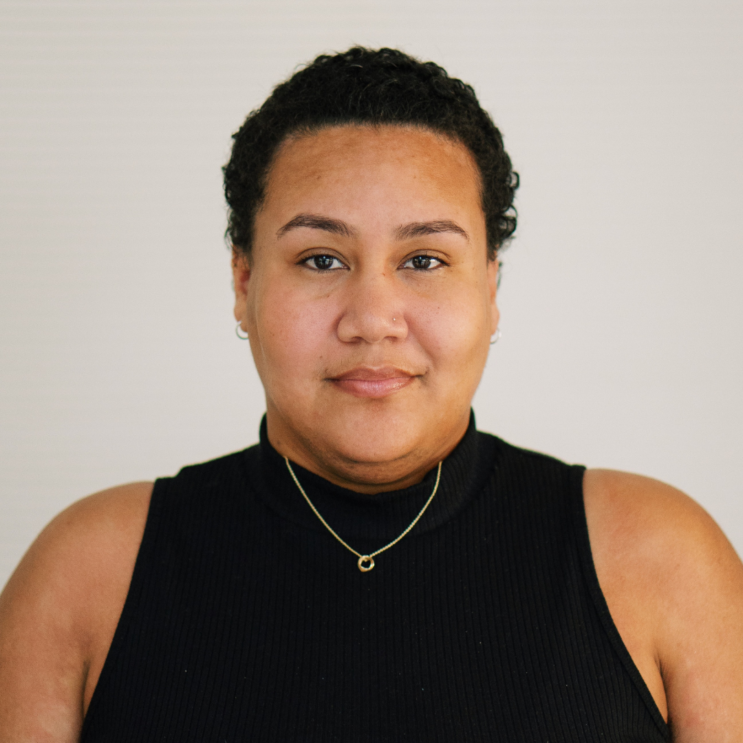 A Black person in a black mock-neck tank top and a thin gold necklace. Their hair is cropped short and they gaze straight ahead.