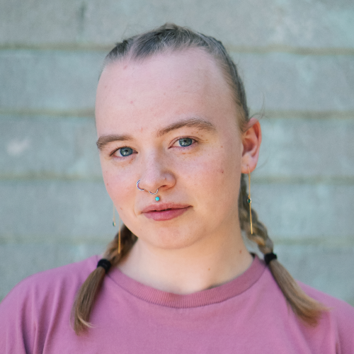A young person with fair skin, blue eyes, and freckles wearing a mauve t-shirt, a septum ring, and earrings, standing in front of a gray brick wall.