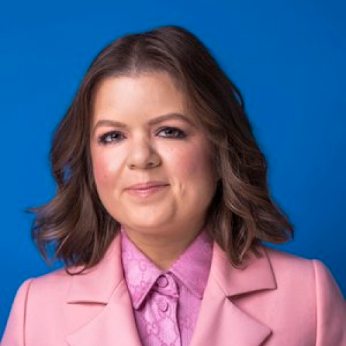 Portrait of a woman with shoulder-length brown hair, wearing a pink blazer and a pink patterned blouse, standing against a blue background.