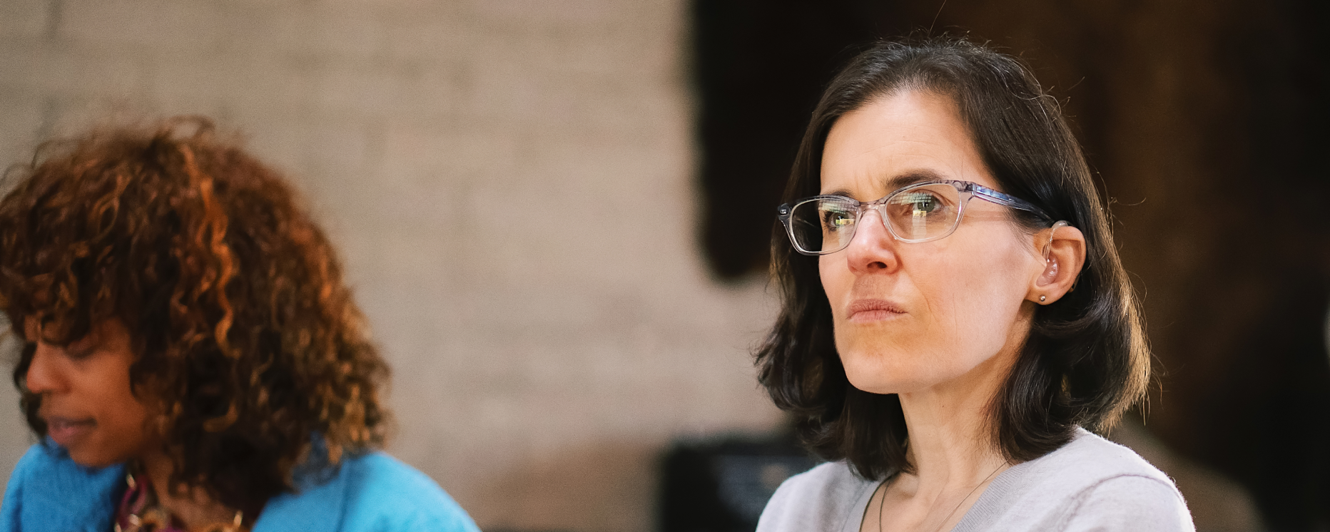 A woman with black hair, wearing glasses and a gray shirt, looking to the right with a serious expression. A woman with curly hair and a blue shirt is partially visible on the left.