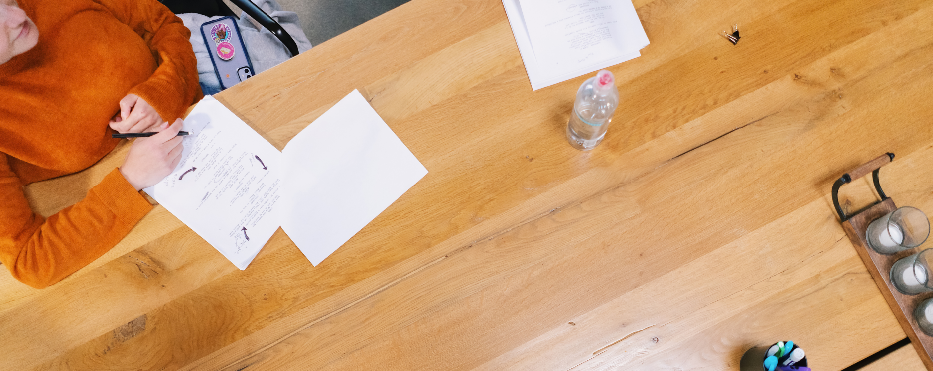 Top-down view of a wooden table with a person in an orange sweater writing on a paper, a water bottle, a pair of glasses, a clip, and some candle holders.