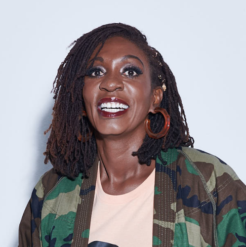 A woman with dark skin, styled dreadlocks, bright makeup, and large hoop earrings smiling at the camera. She is wearing a camouflage jacket over a light-colored top, against a plain background.