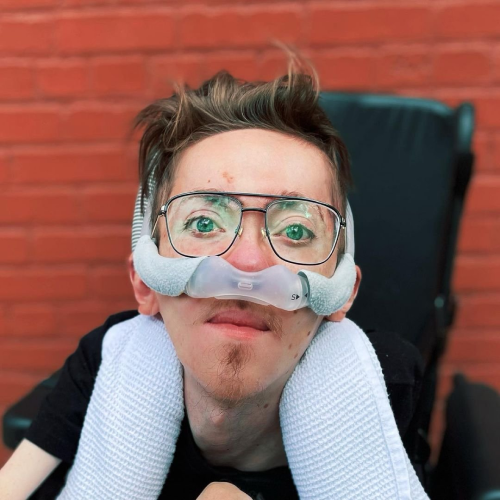 A man with glasses and facial hair using a nasal mask, resting their face on two rolled towels, with a brick wall in the background.