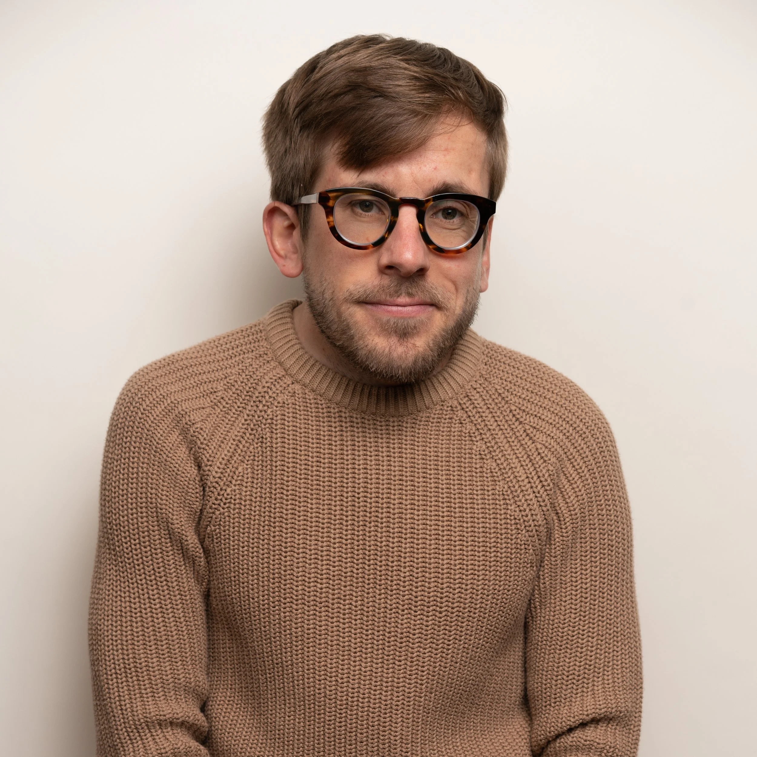 A man with short brown hair, wearing large tortoiseshell glasses and a tan knit sweater, smiling slightly against a plain white wall.
