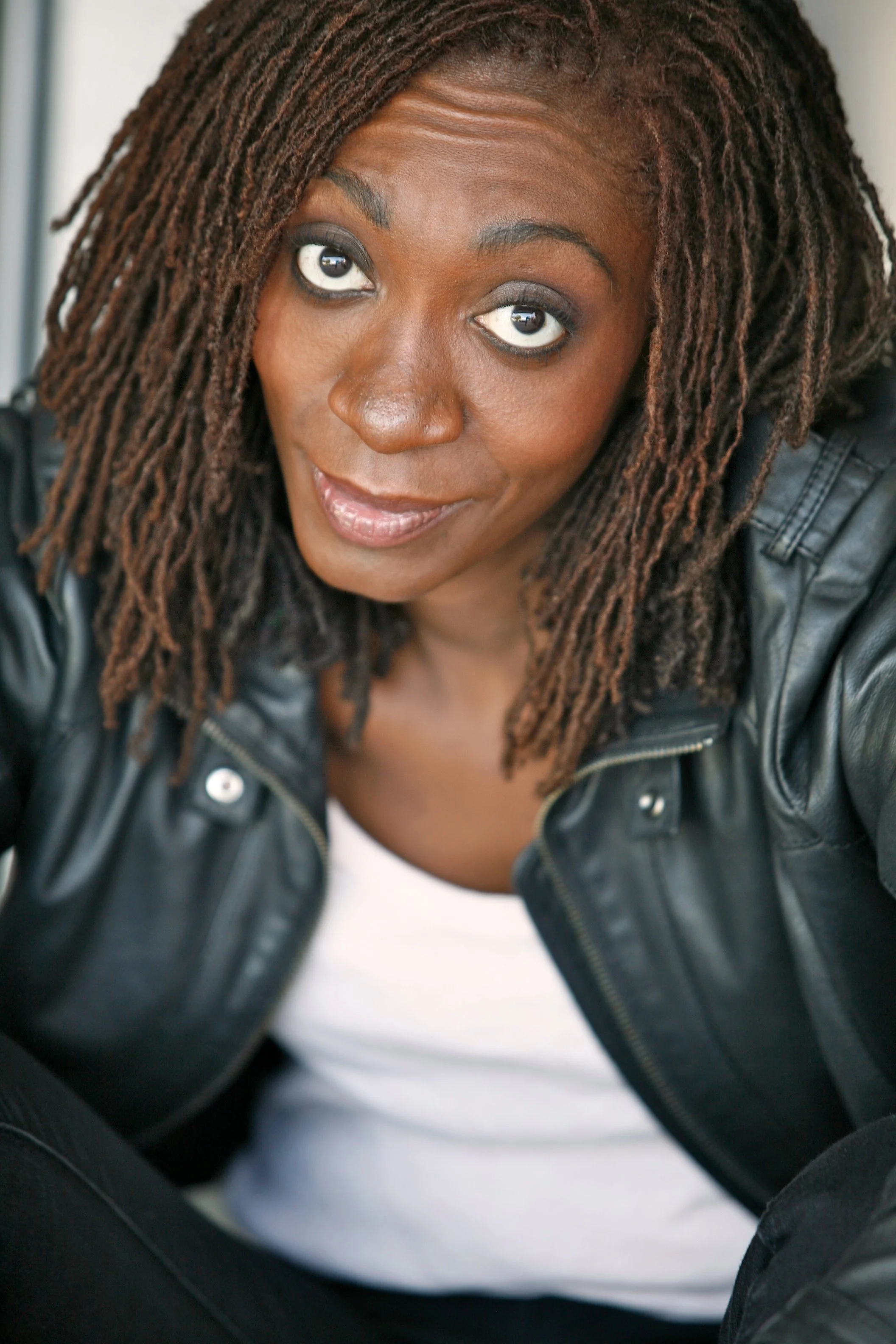Close-up of a woman with long, brown dreadlocks, wearing a black leather jacket and a white shirt, looking upward with a slight smile.
