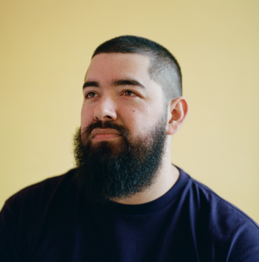 A man with short dark hair, a full beard, and a mustache, wearing a navy blue shirt, looking slightly to the right against a yellow background.