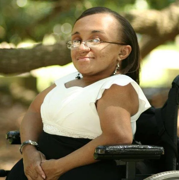 A woman with dark hair, glasses, and earrings sitting outdoors on a bench with a blurred background of trees.
