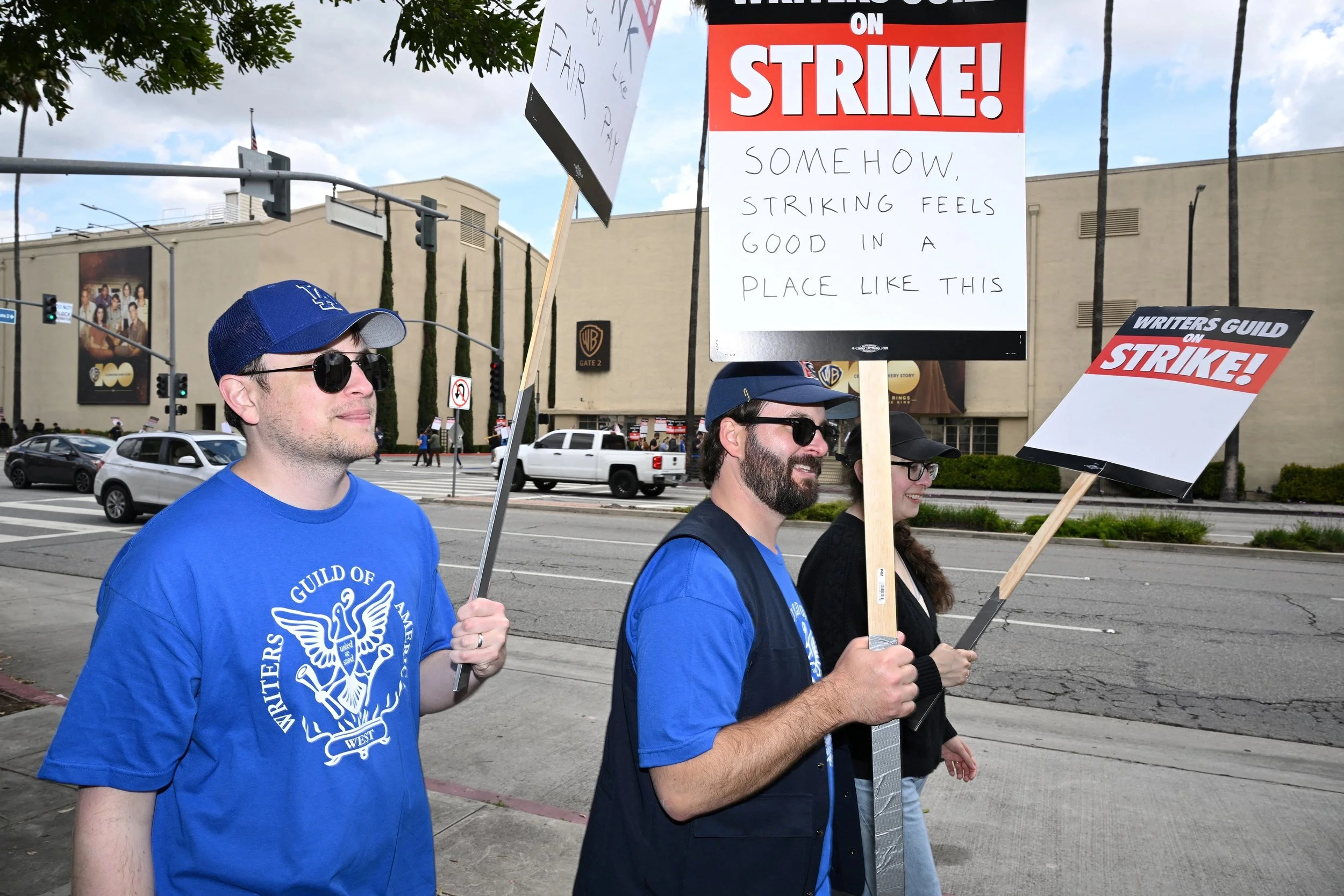 Three people wearing baseball caps, sunglasses and blue t-shirts that say Writers Guild or Americas with the WGA logo walking by the side of a busy street and holding picket signs that read, “Writers Guild on Strike! Somehow striking feels good in a