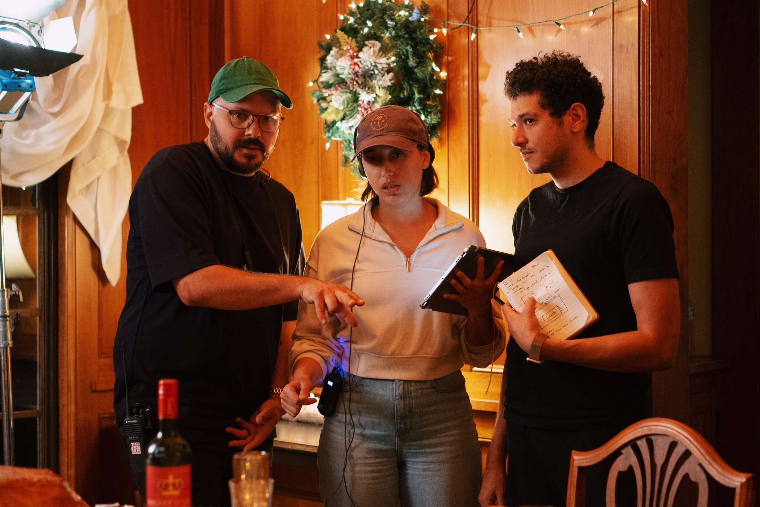 Three people stand on a christmas-decorated set in wrapt conversation. They are looking at the same place on a table in front of them, and using their hands to illustrate what they're discussing.