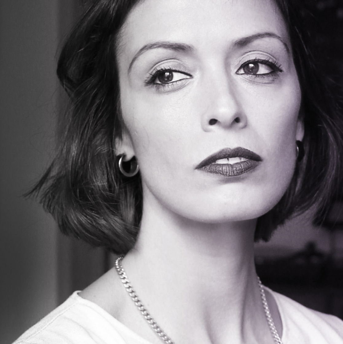 A black and white headshot of a person with dark hair in a bob wearing dark lipstick, a silver chain necklace and a white top.
