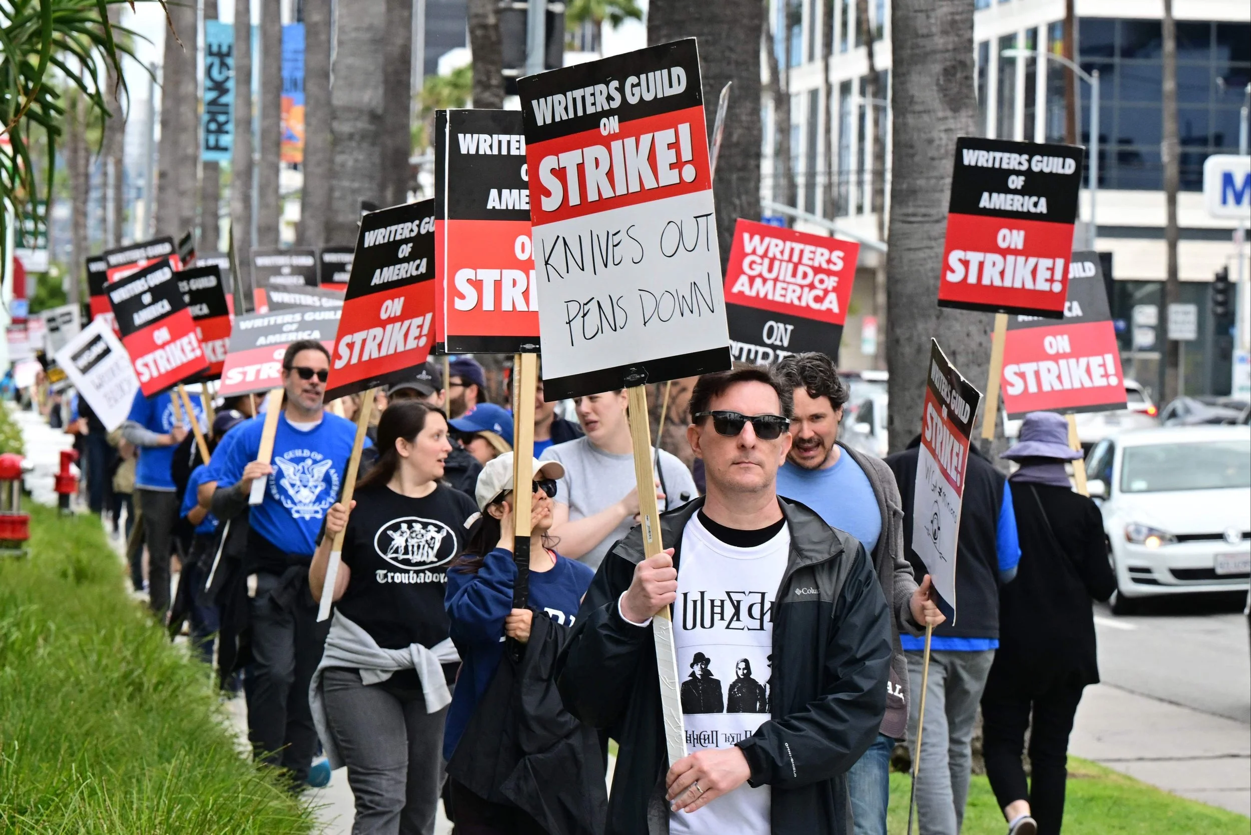 A large group of people with walking on a palm-tree lined sidewalk with picket signs that say "Writers Guild on Strike!"