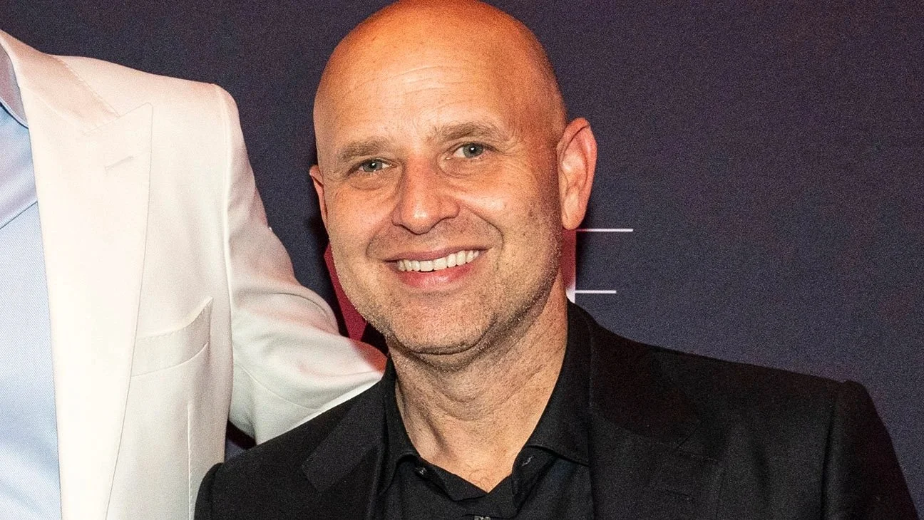 Image Description: A photo of Ari Greenburg, a bald man with blue eyes, smiling in front of a step and repeat and wearing a black suit.