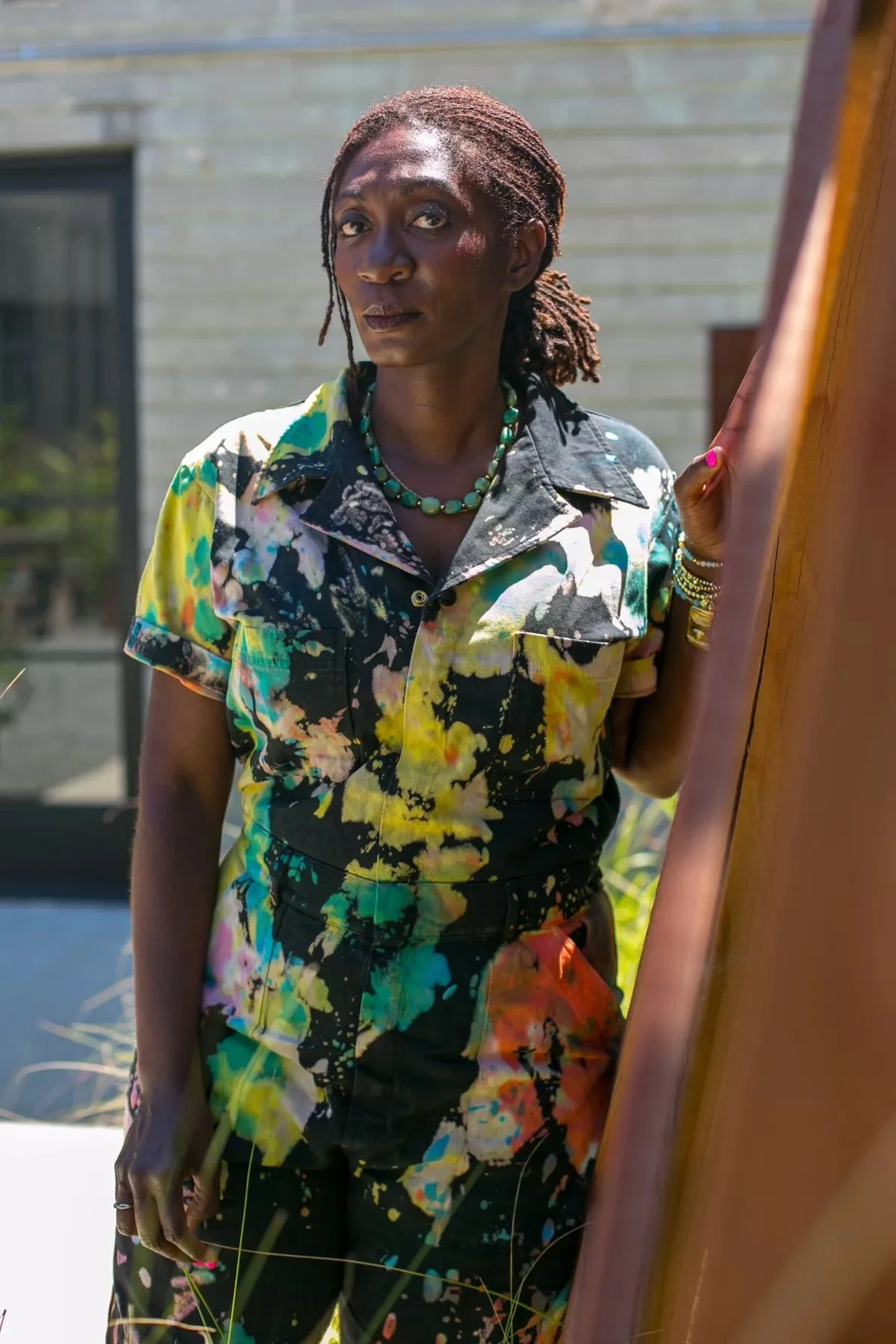 A Black woman stands outside and looks into the camera with her hair in a ponytail. She wears a tie-dye multicolored jumpsuit and a necklace with big turquoise stones.