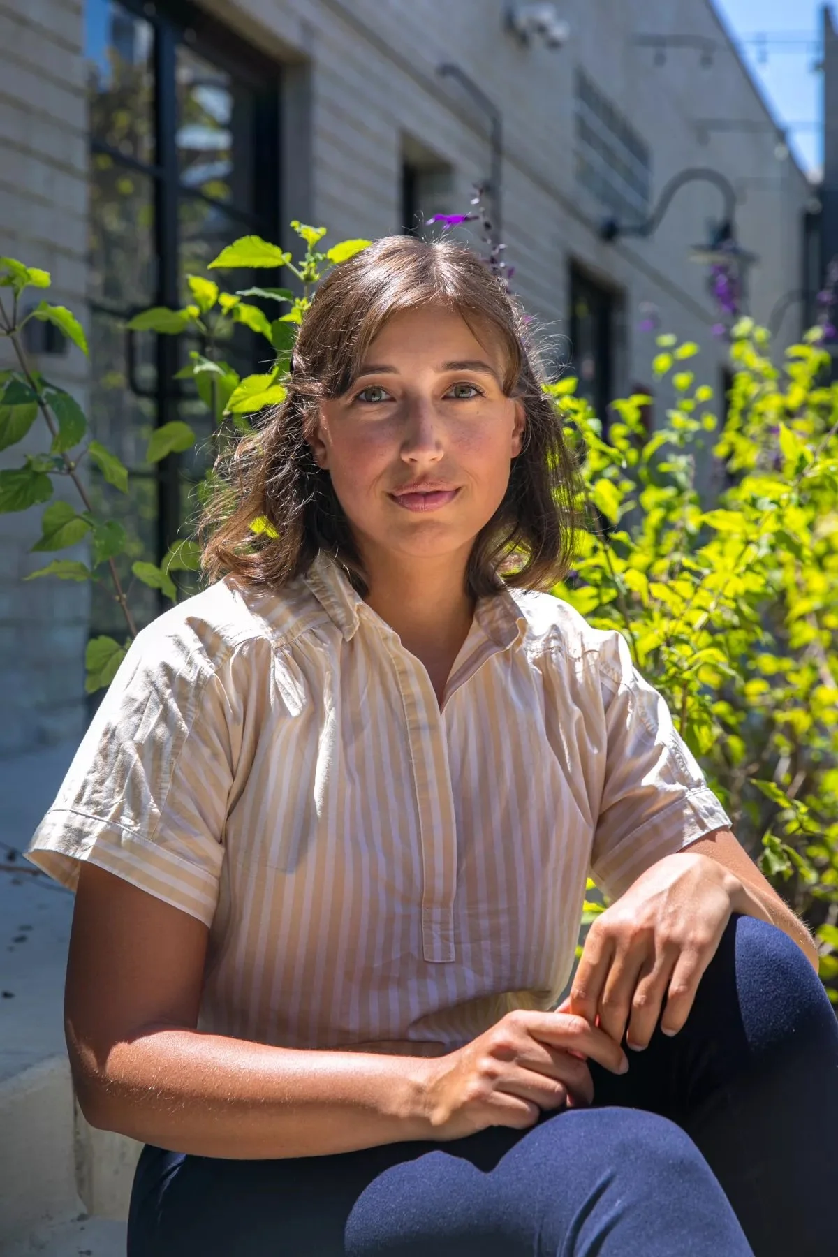 A woman with shoulder-length brown hair sits and looks into the camera. She wears a white and beige striped shirt and blue jeans. Behind her are tall plants and flowers.