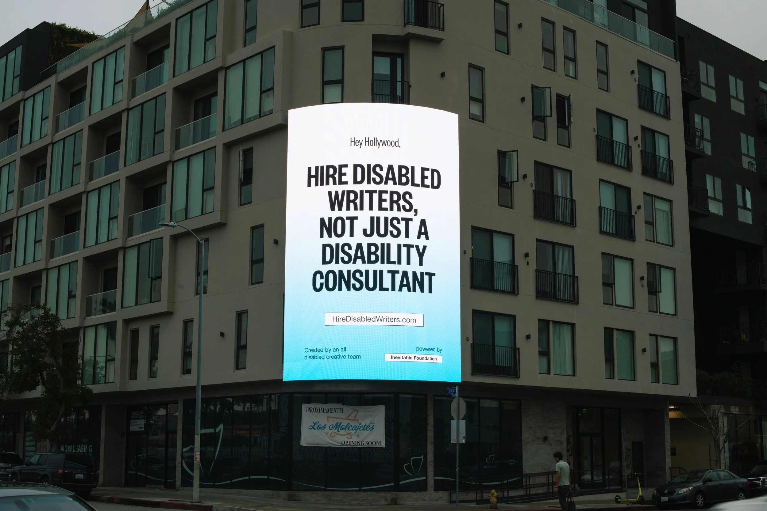A photograph of a billboard against a three story building, lit up with a blue and white gradient background and bold black text that reads: “Hey Hollywood, Hire Disabled Writers, Not Just A Disability Consultant”