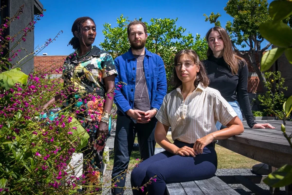An image of four people outside surrounded by flowers, looking directly into camera.
