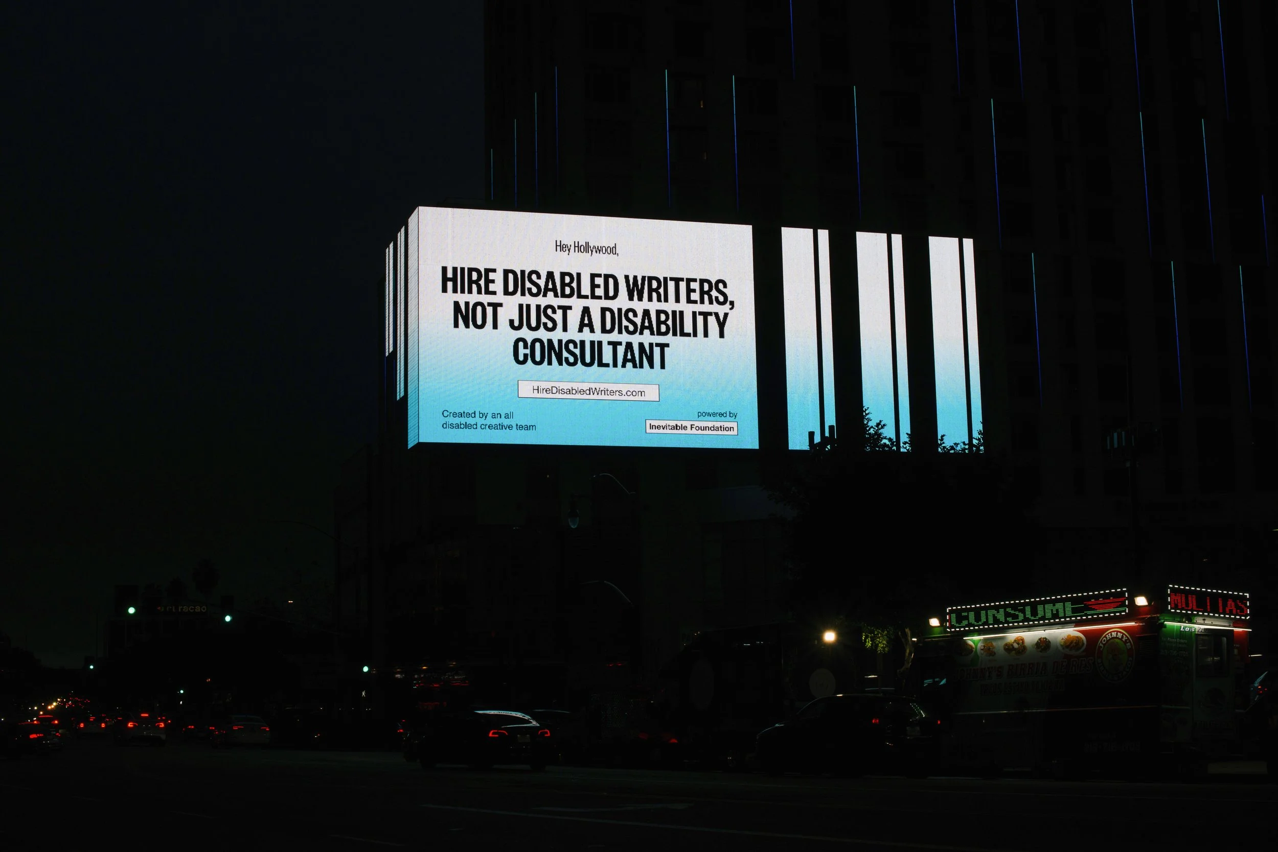 A large billboard lit up at night over a busy street. It shows bold black text over a blue and white gradient background that reads: "Hey Hollywood, Hire Disabled Writers, Not Just A Disability Consultant"