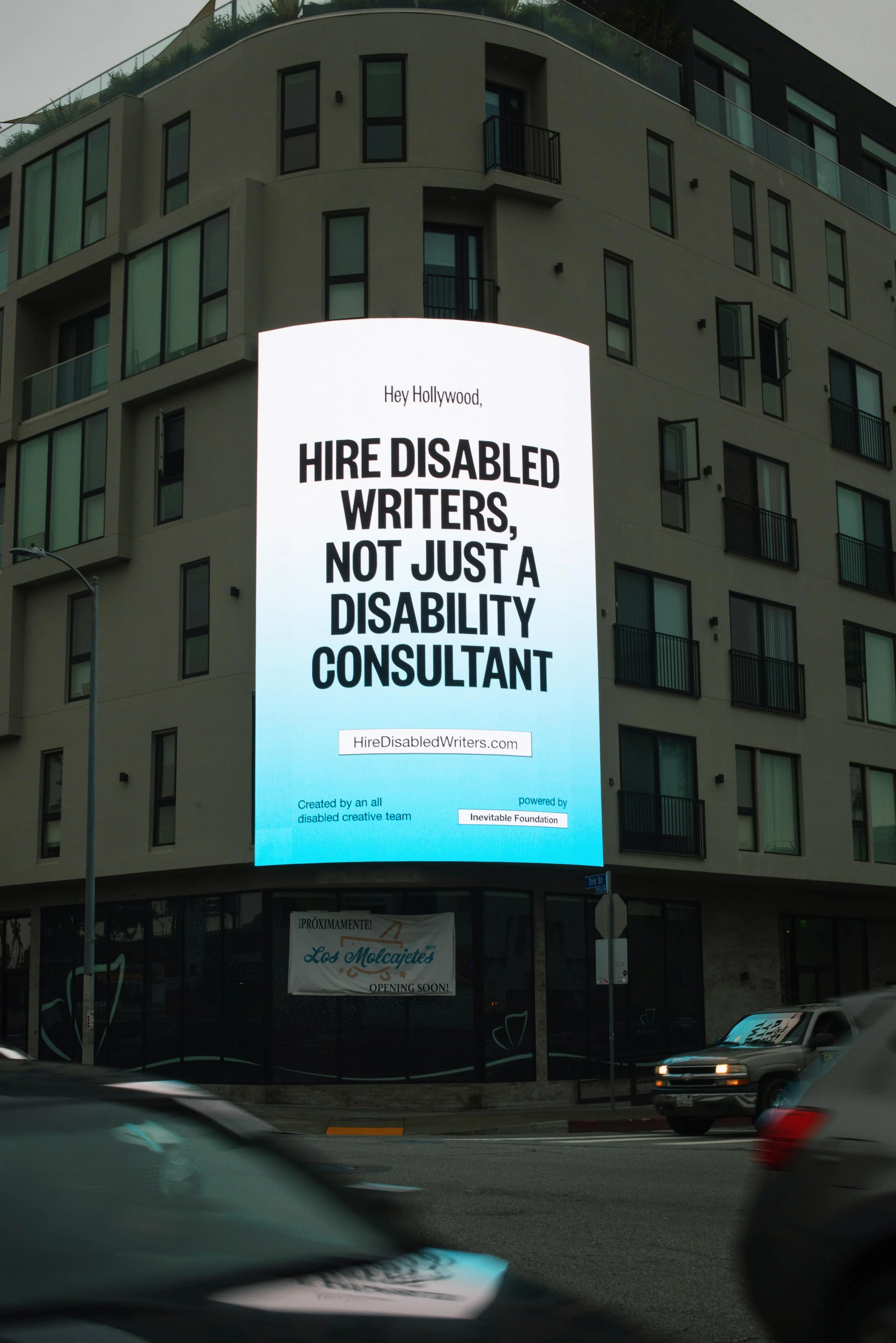 A three-story billboard lit up on the side of a building. It shows bold black text against a blue and white gradient background that reads "Hey Hollywood, Hire Disabled Writers, Not Just A Disability Consultant."