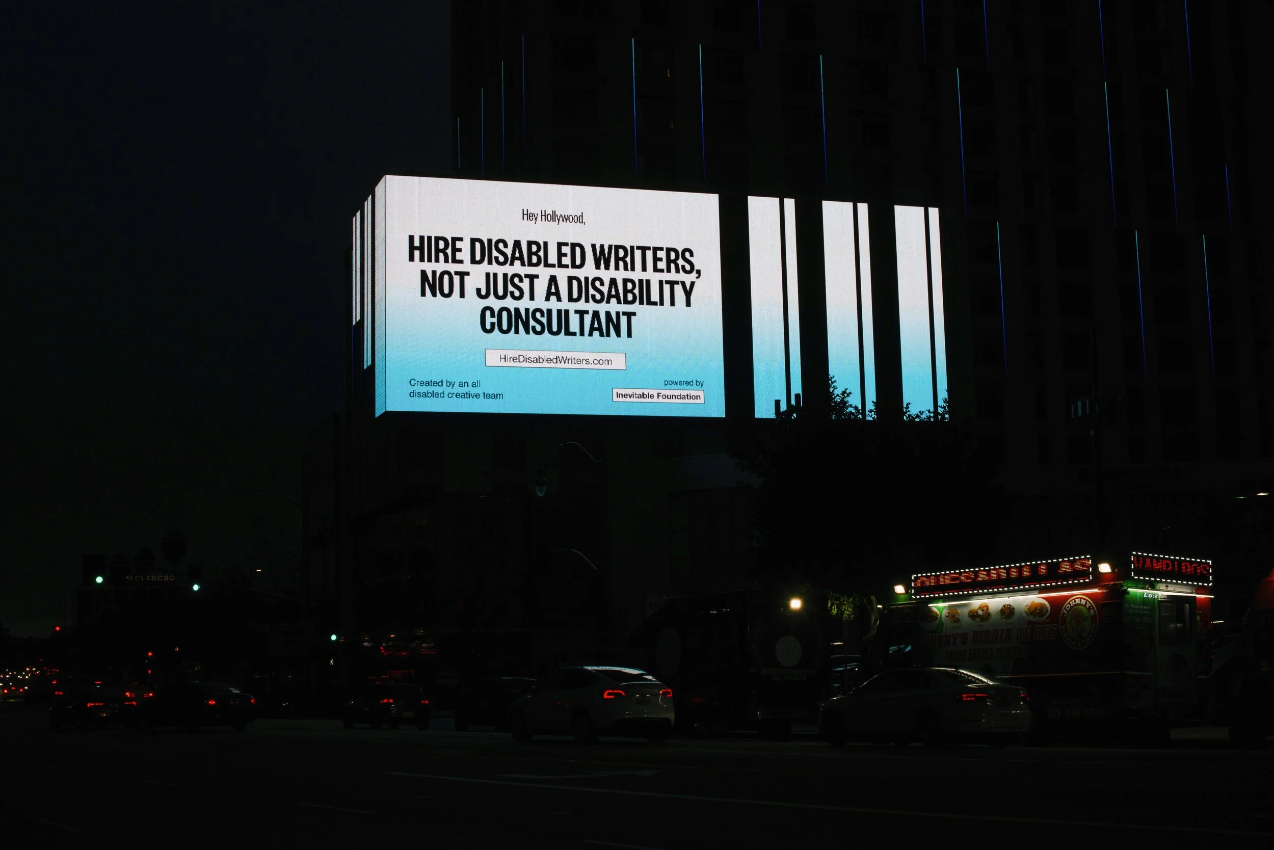 A large billboard lit up at night over a busy street. It shows bold black text over a blue and white gradient background that reads: "Hey Hollywood, Hire Disabled Writers, Not Just A Disability Consultant"