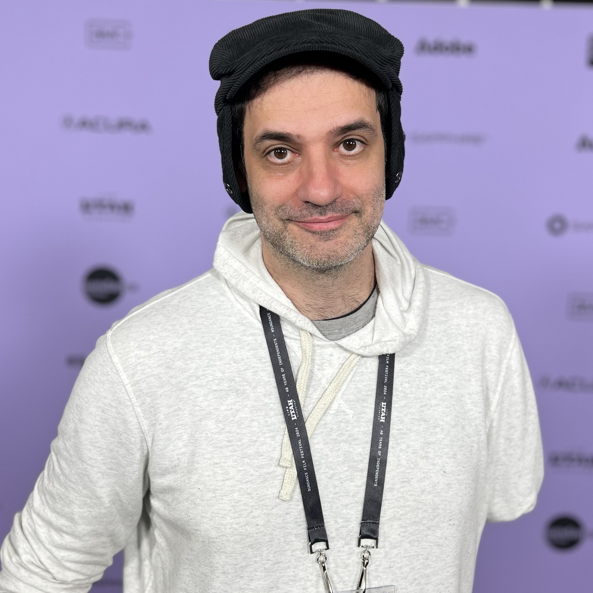 A Hispanic man with brown eyes wearing a blue earflap hat, a white jumper, and a film festival badge, with a missing left arm, and smiling.