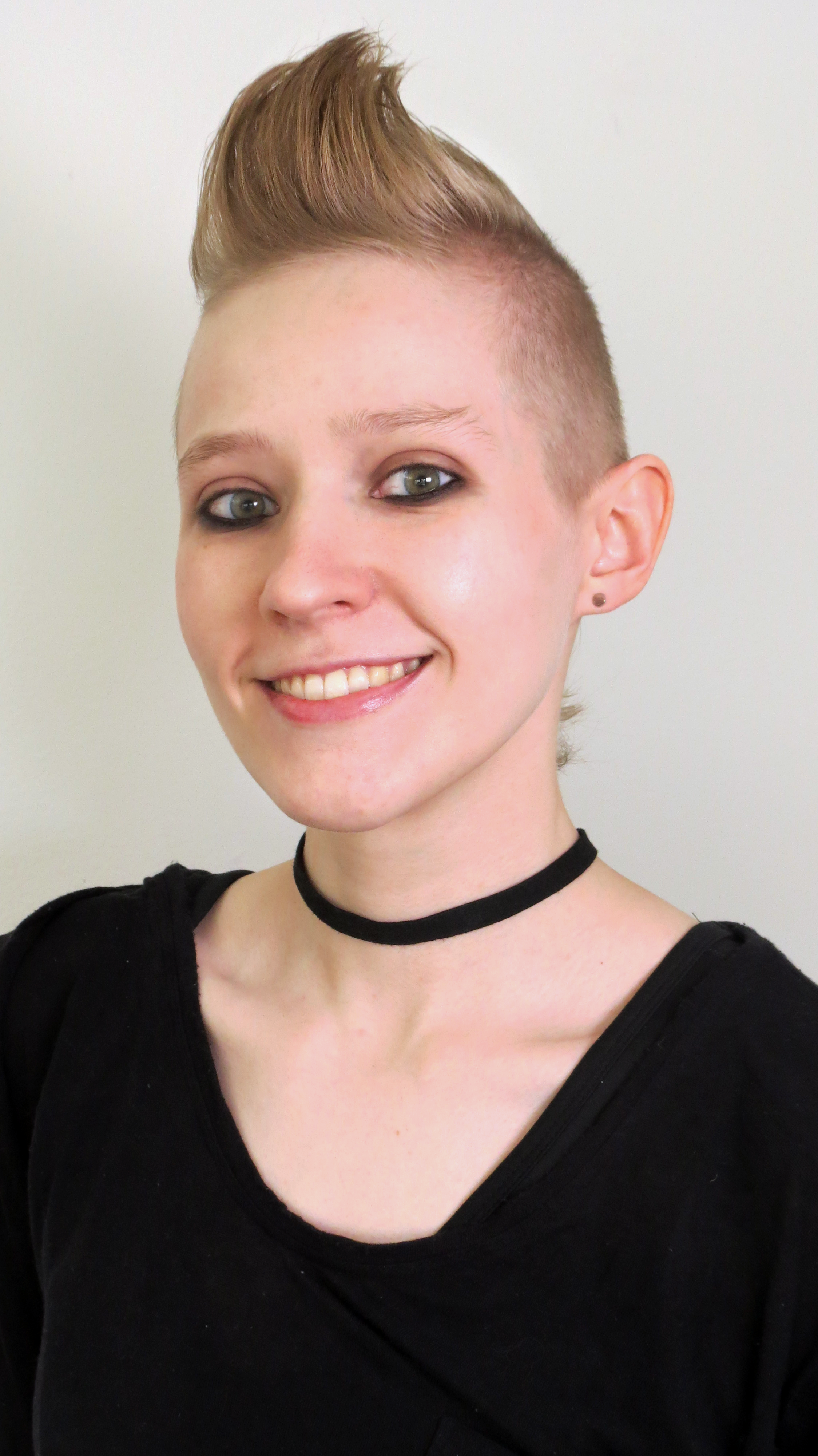 A white woman in her mid 20’s with a blonde mohawk. She’s wearing a black shirt, a black necklace, and grinning at the camera.