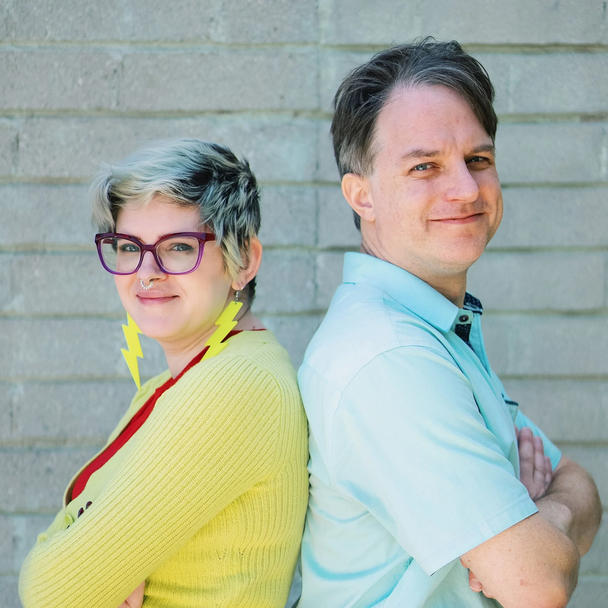 Two people standing back-to-back. The person on the left has short light hair, wears bright yellow glasses, and is dressed in a bright yellow sweater. The person on the right has short brown hair and is wearing a light blue collared shirt.