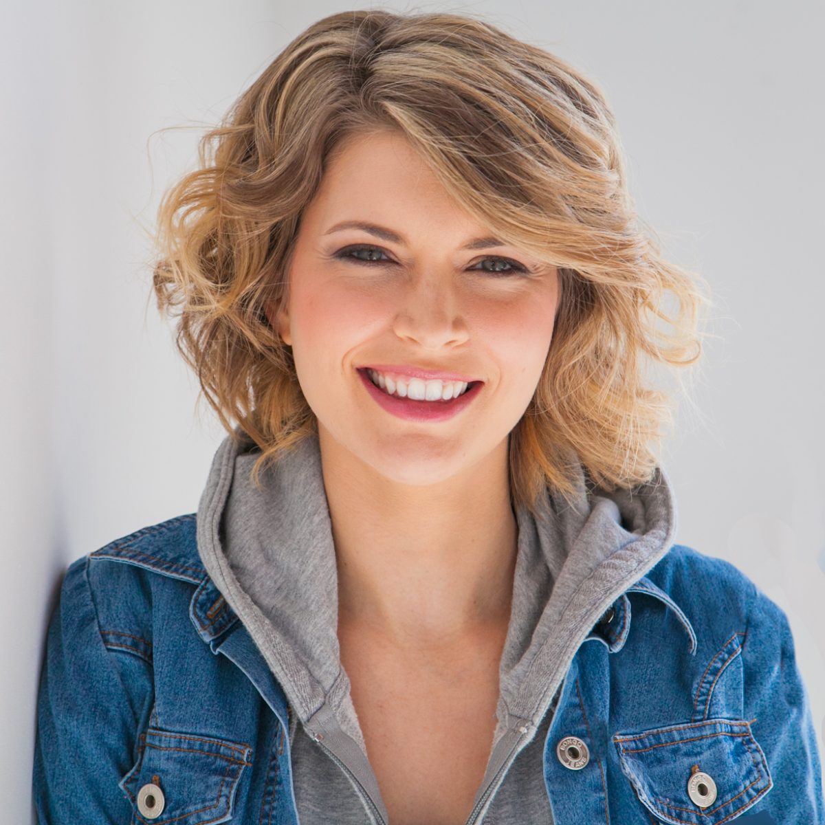 A caucasian woman with wavy blond hair and blue eyes, smiling at the camera. She is wearing a blue jean jacket and is in front of a white background.