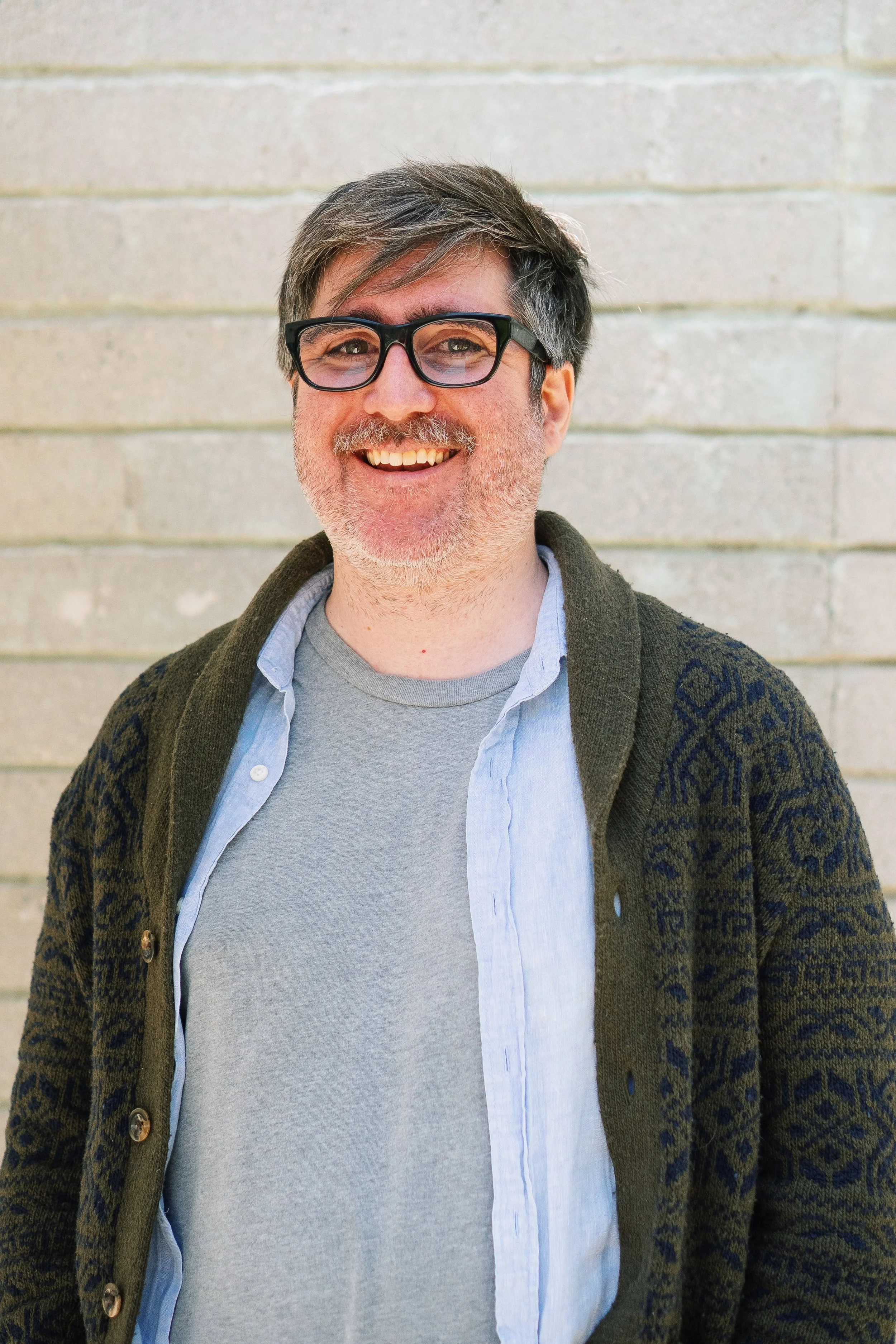 A headshot of a smiling man with salt and pepper short hair, black thick-framed glasses and a light mustache and beard. He is wearing a grey t-shirt with a blue button-up shit and grey and blue patterned cardigan.